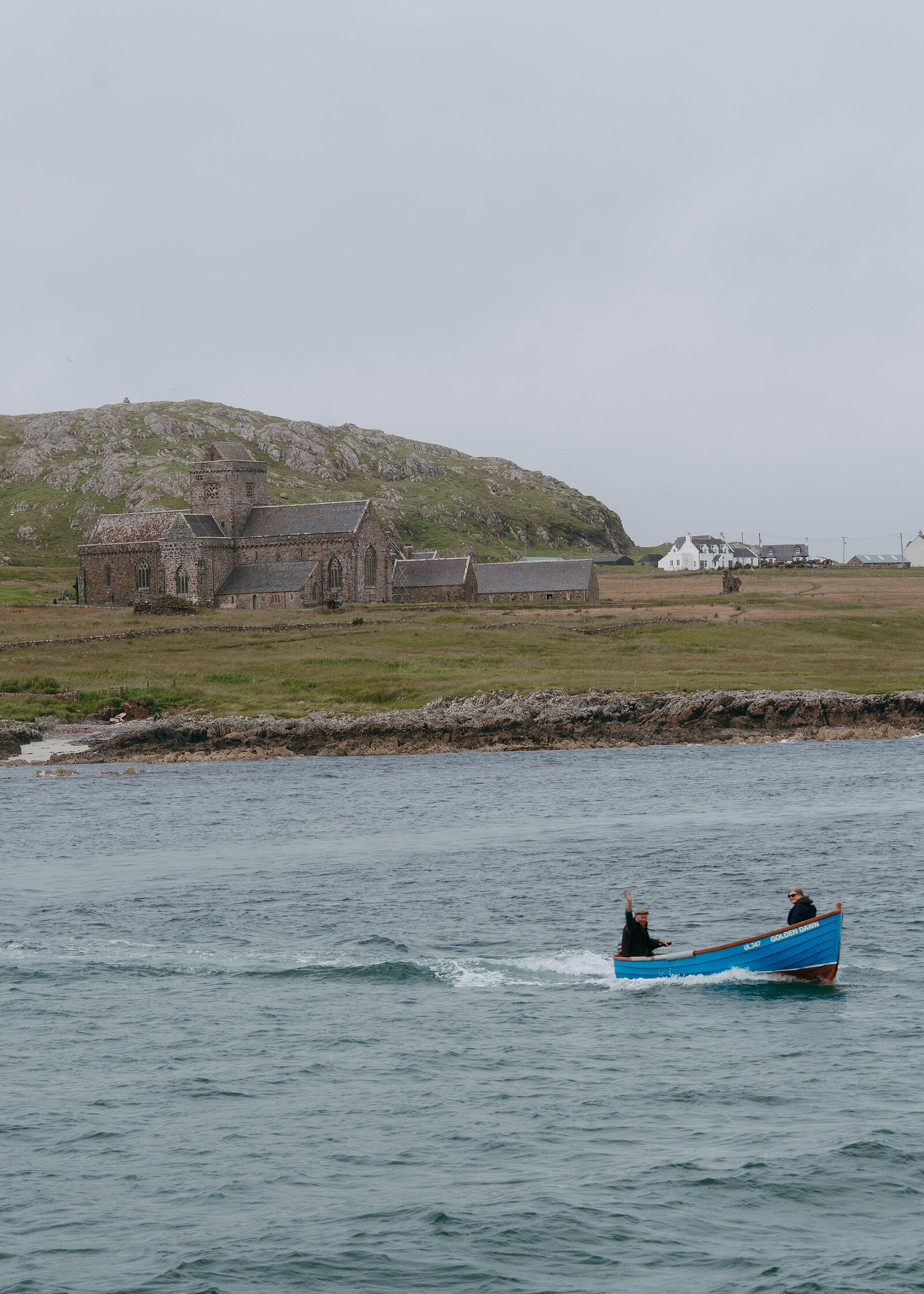 Closer on the blue rowing boat and its two passengers passing Iona Abbey, white houses along the shore.