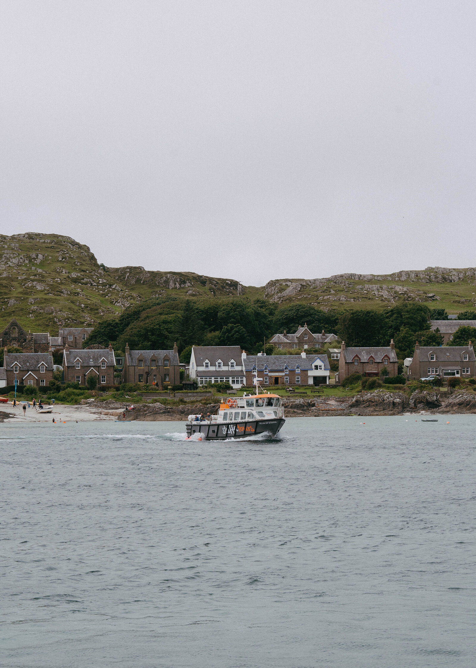 A small passenger boat leaving the Iona shore, a row of stone houses and a green hill behind.