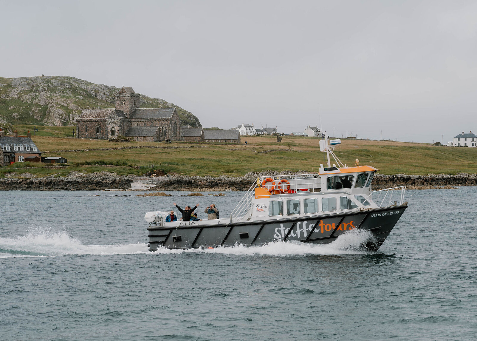 The Staffa Tours boat speeding past Iona Abbey, passengers waving from the back deck.