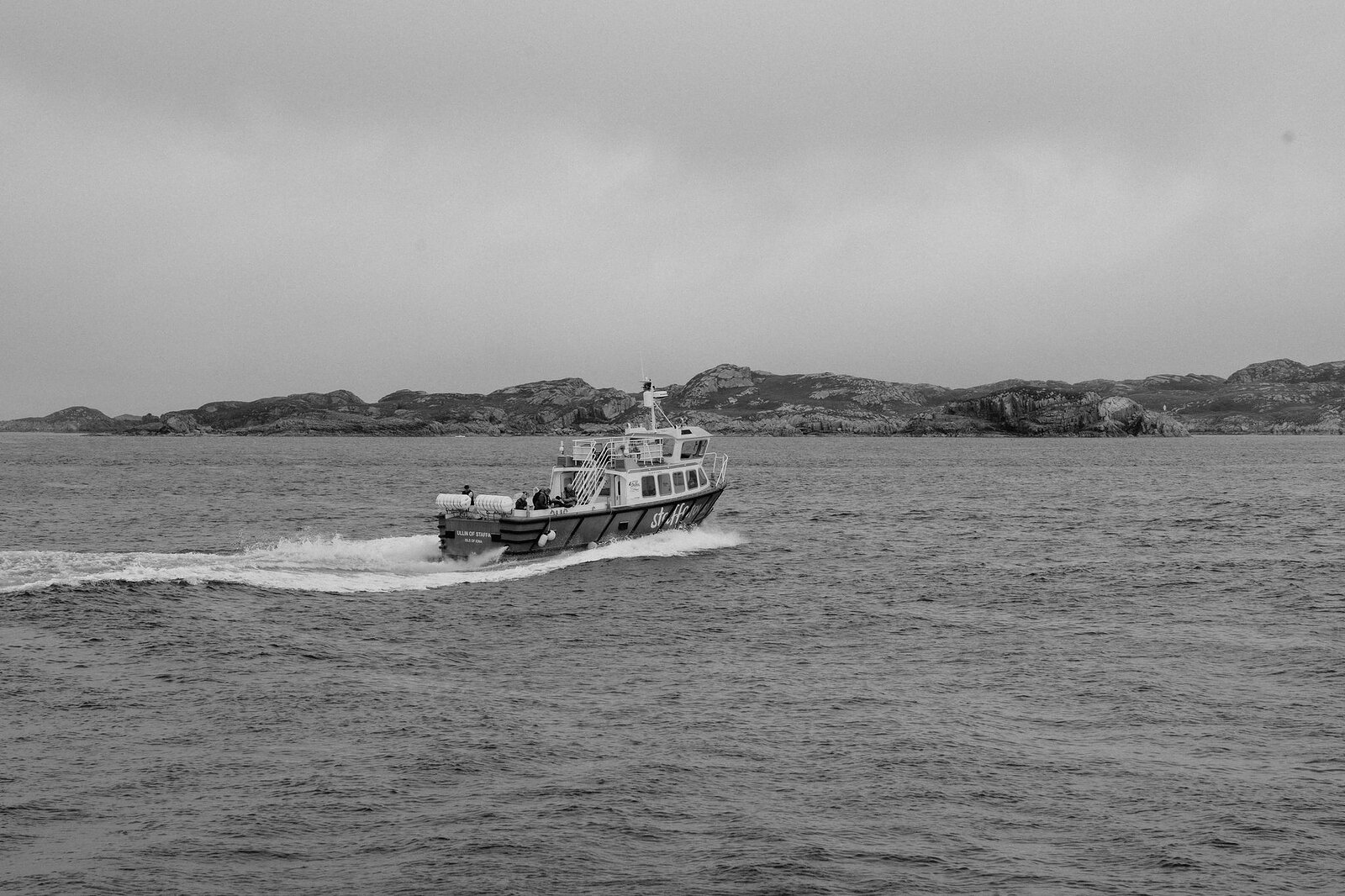 A black-and-white frame of the tour boat throwing up a long wake across the sound, low islands behind.