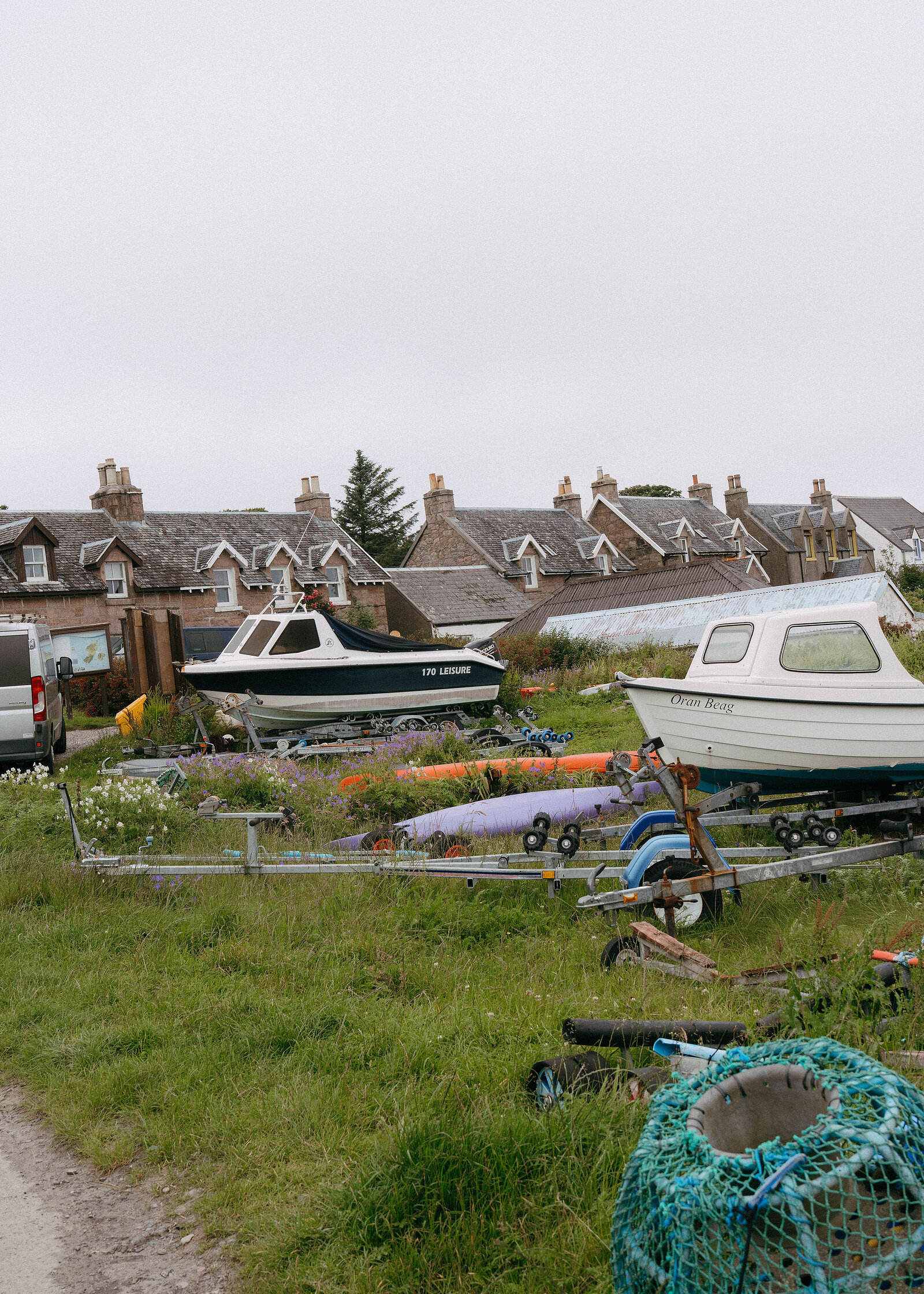 Boats on trailers and a lobster pot in the grass above the shore, a row of village houses behind.
