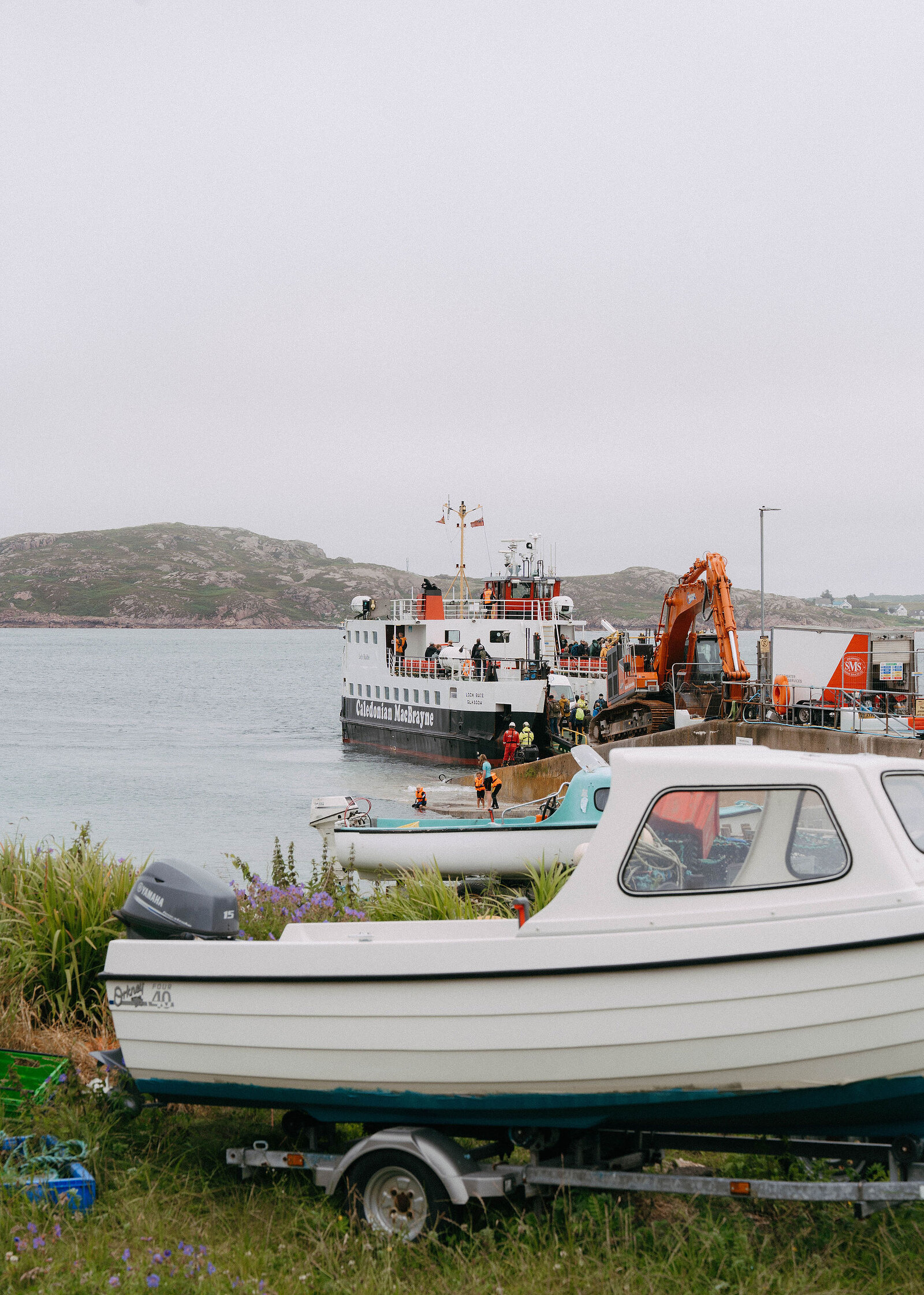 A white boat on a trailer in the grass, the CalMac ferry loading at the slipway beside an orange digger.