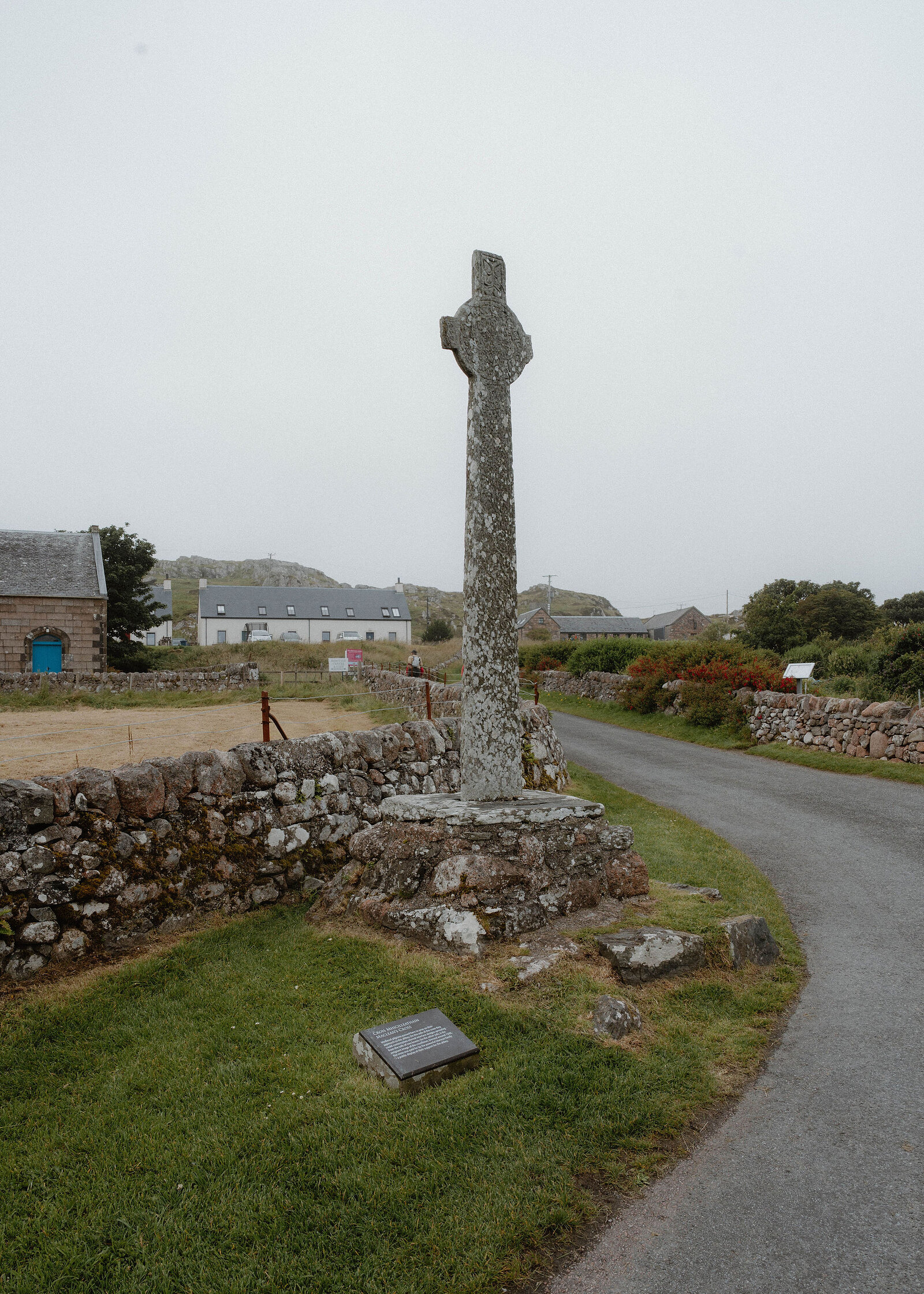 A tall carved stone cross beside a single-track road and a dry-stone wall, white cottages behind.
