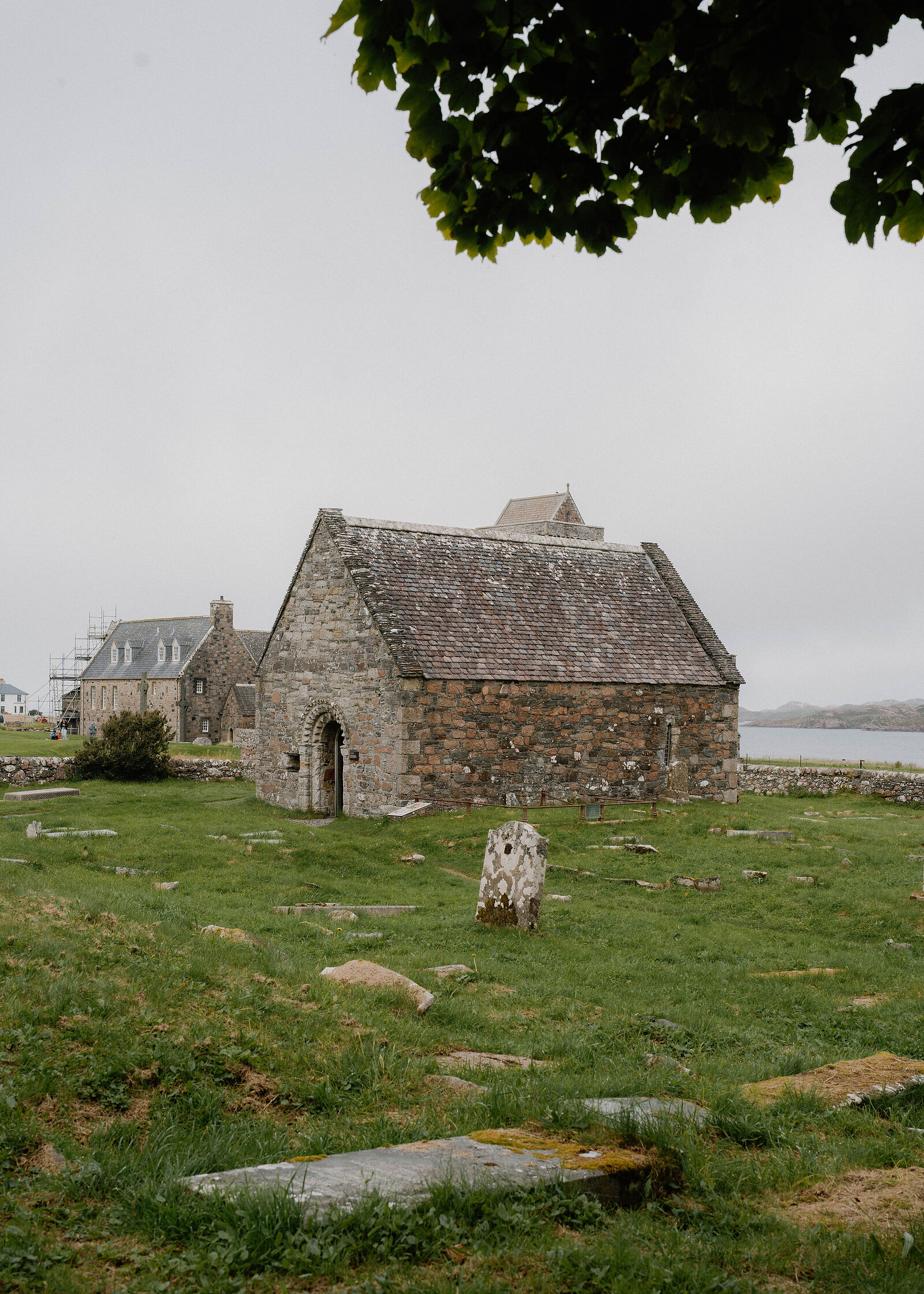 A small stone chapel with a slate roof in a grassy graveyard, the abbey buildings and sea behind.