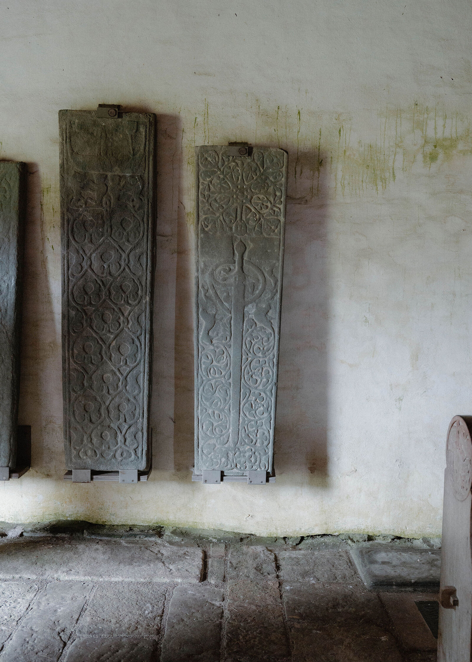 Two carved medieval grave slabs standing against the chapel's whitewashed wall above a flagstone floor.