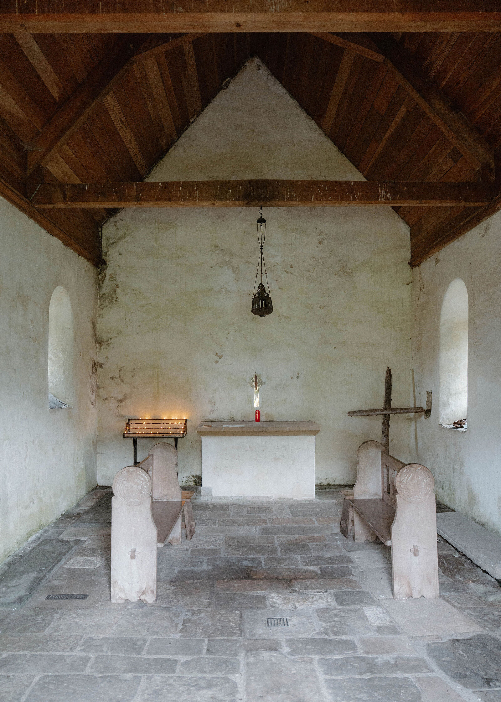 Inside the chapel: a timber roof, a plain altar with a candle, simple benches and a flagstone floor.