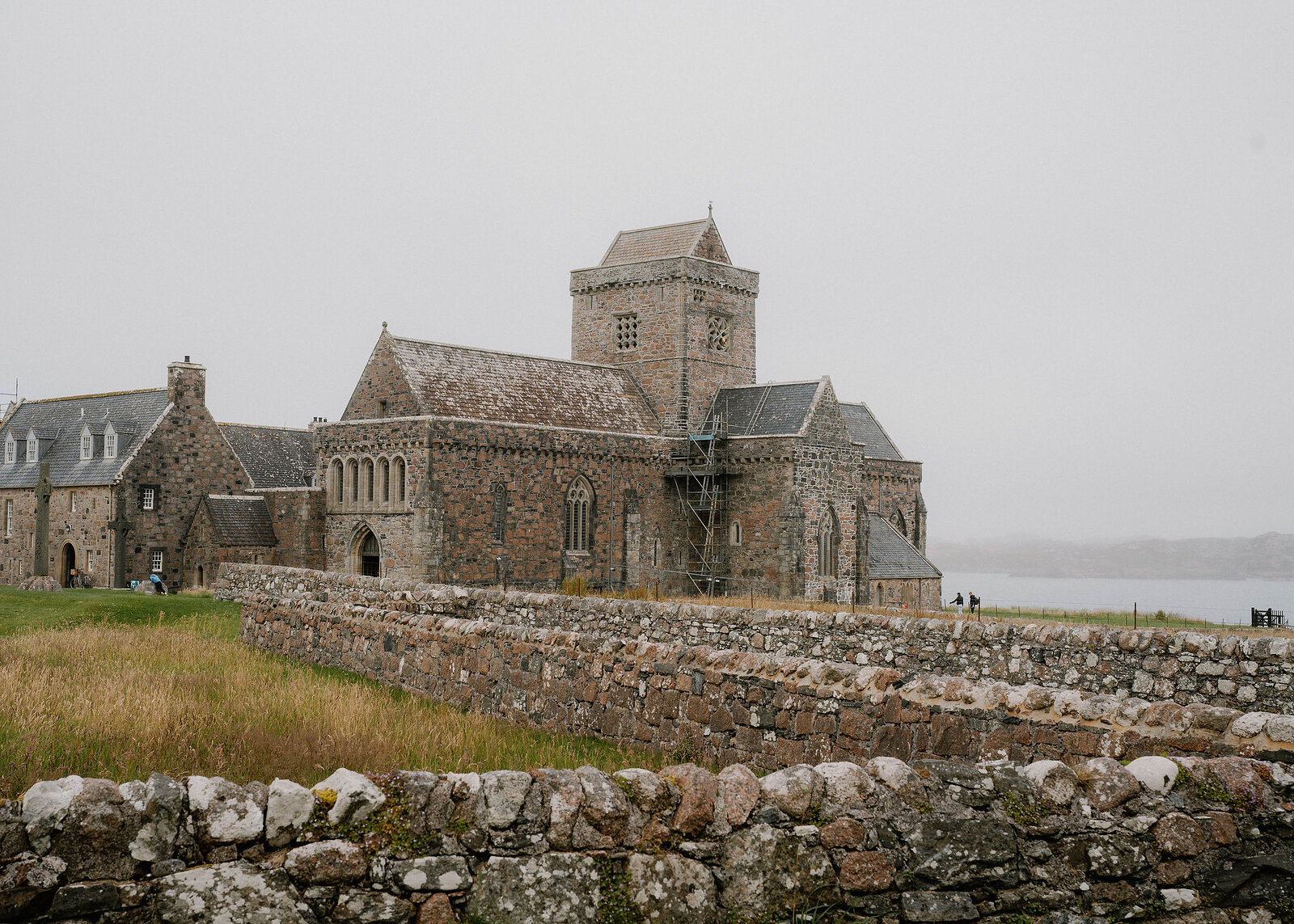 A landscape view of Iona Abbey above lichened stone walls, the sound grey behind and a few visitors by the door.