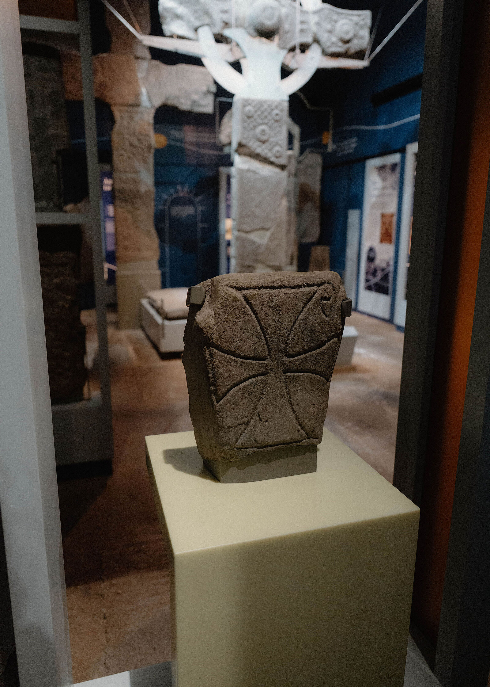A carved stone with a cross on a pale plinth in the abbey museum, a tall cross and panels behind.