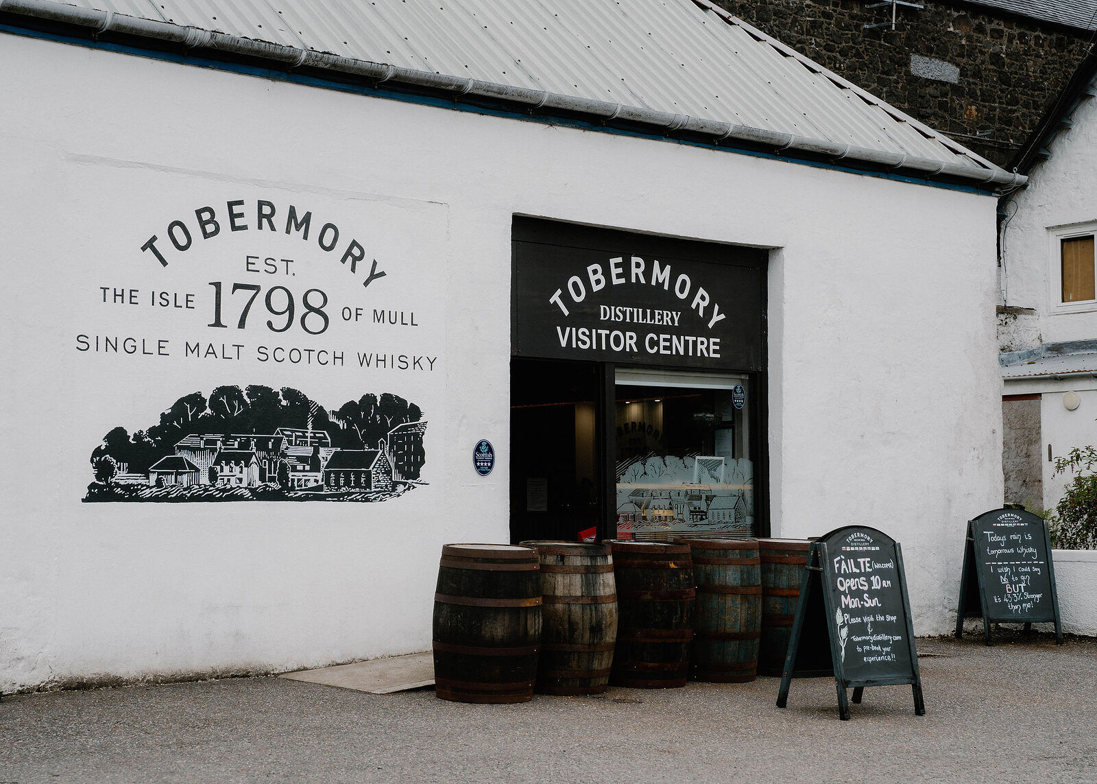 A landscape view of the distillery's whitewashed wall painted 'Tobermory, est. 1798', barrels by the visitor centre door.