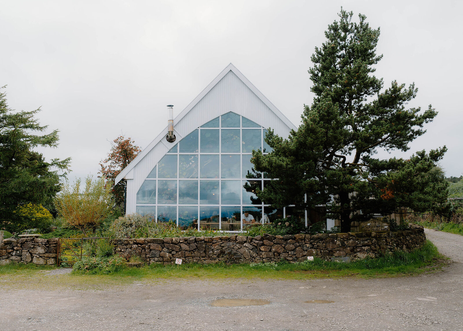 A landscape view of the glass barn's glazed gable, the stone wall and planting in front and pines to each side.