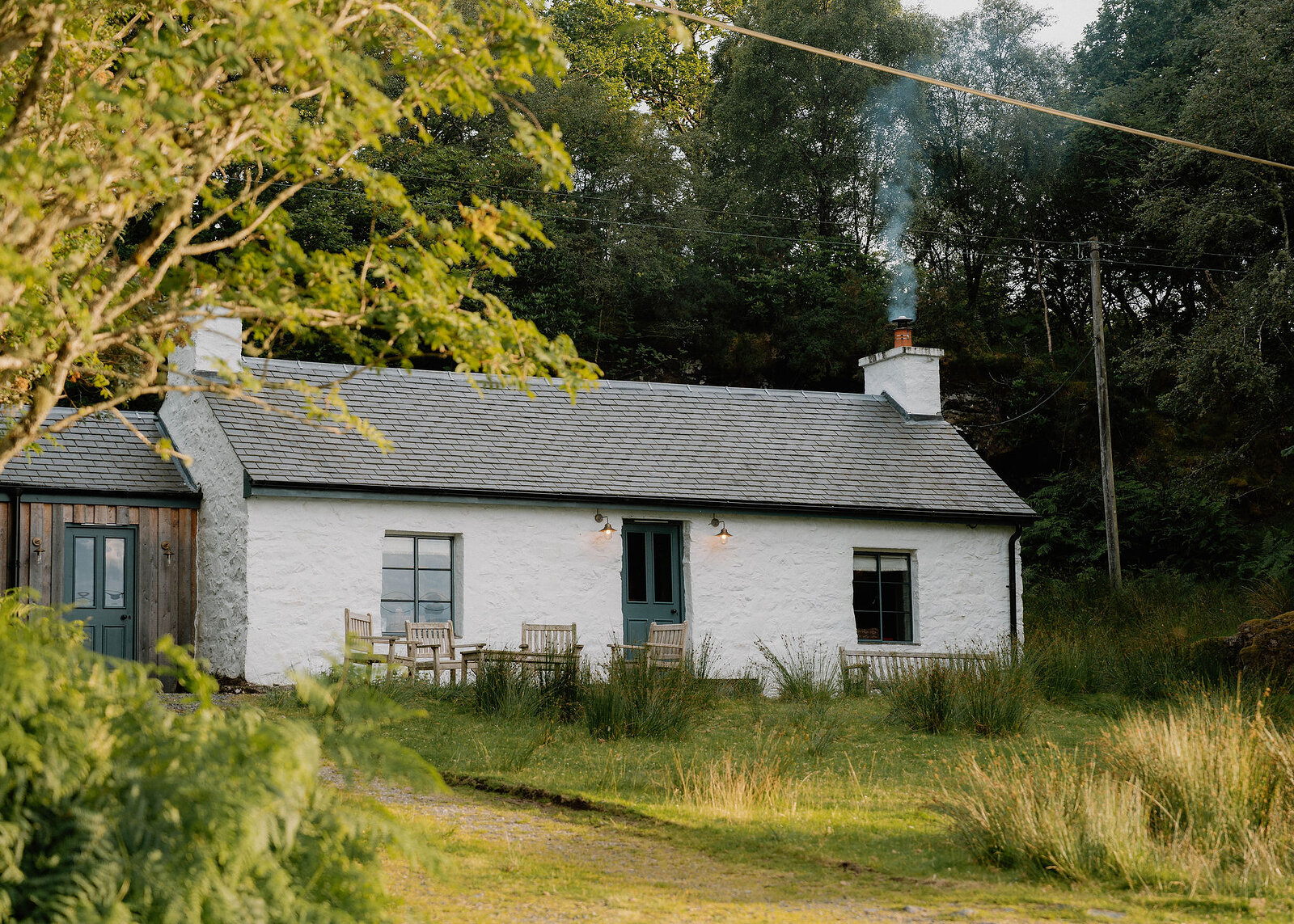 A landscape view of the whitewashed cottage in low evening sun, its blue doors, lit lamps and teak chairs, smoke rising from the chimney.