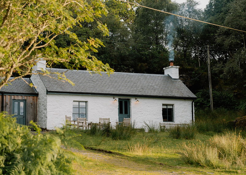 A landscape view of the whitewashed cottage in low evening sun, its blue doors, lit lamps and teak chairs, smoke rising from the chimney.