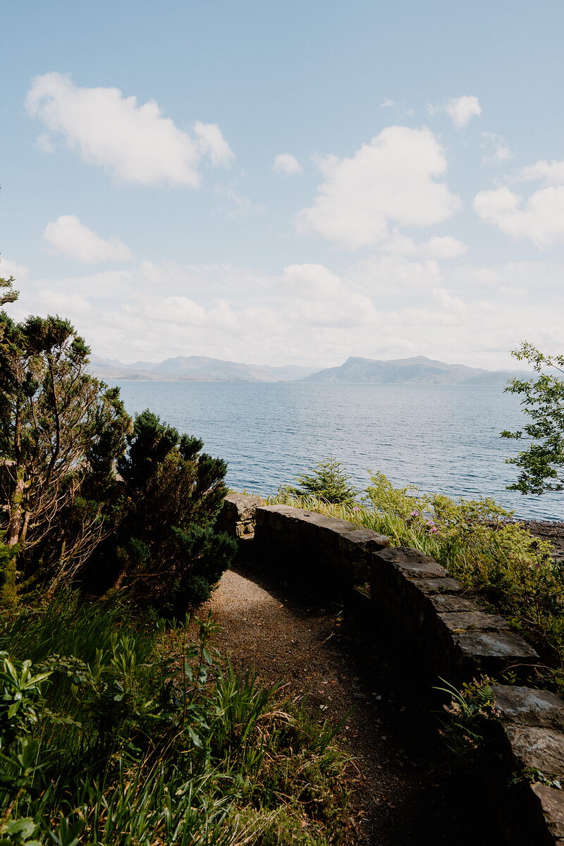 A stone path curves along a garden wall above the sea, distant mountains beyond.