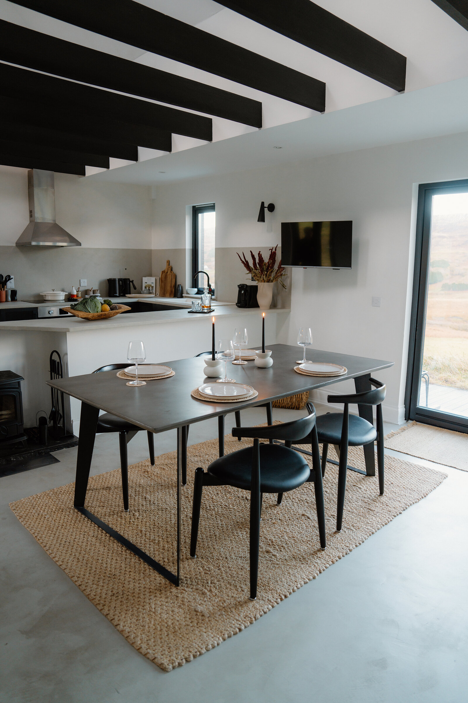A wood burner glows beside a dining table set for a meal, hills visible through glass doors.