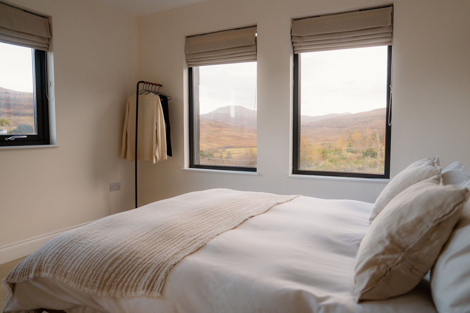 A made-up bed faces windows overlooking hills, a clothes rail standing beside it.