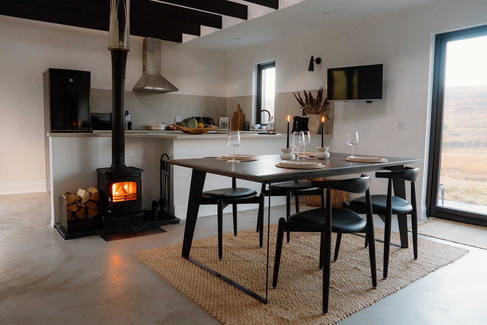 A wood burner glows beside a dining table set with glasses, hills visible through a window.
