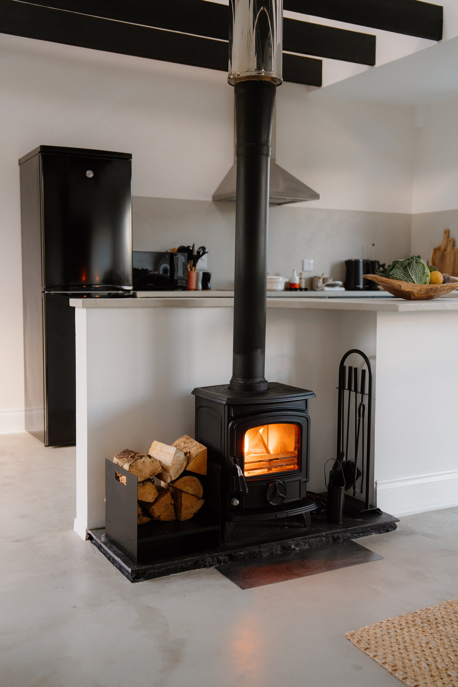 A wood burner glows beside a kitchen counter with a black fridge.