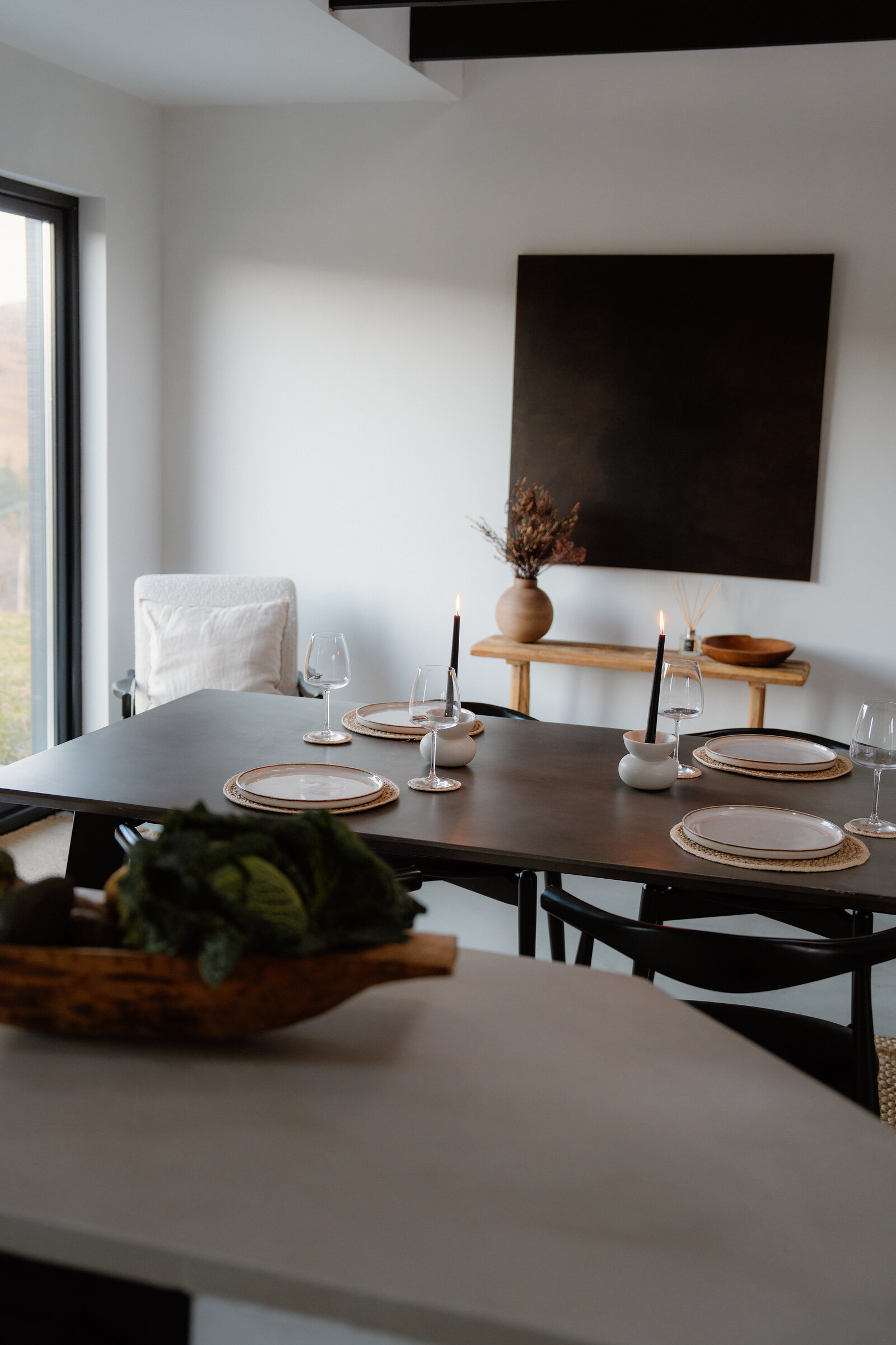 A dining table is set with candles and wine glasses beneath a dark abstract painting.