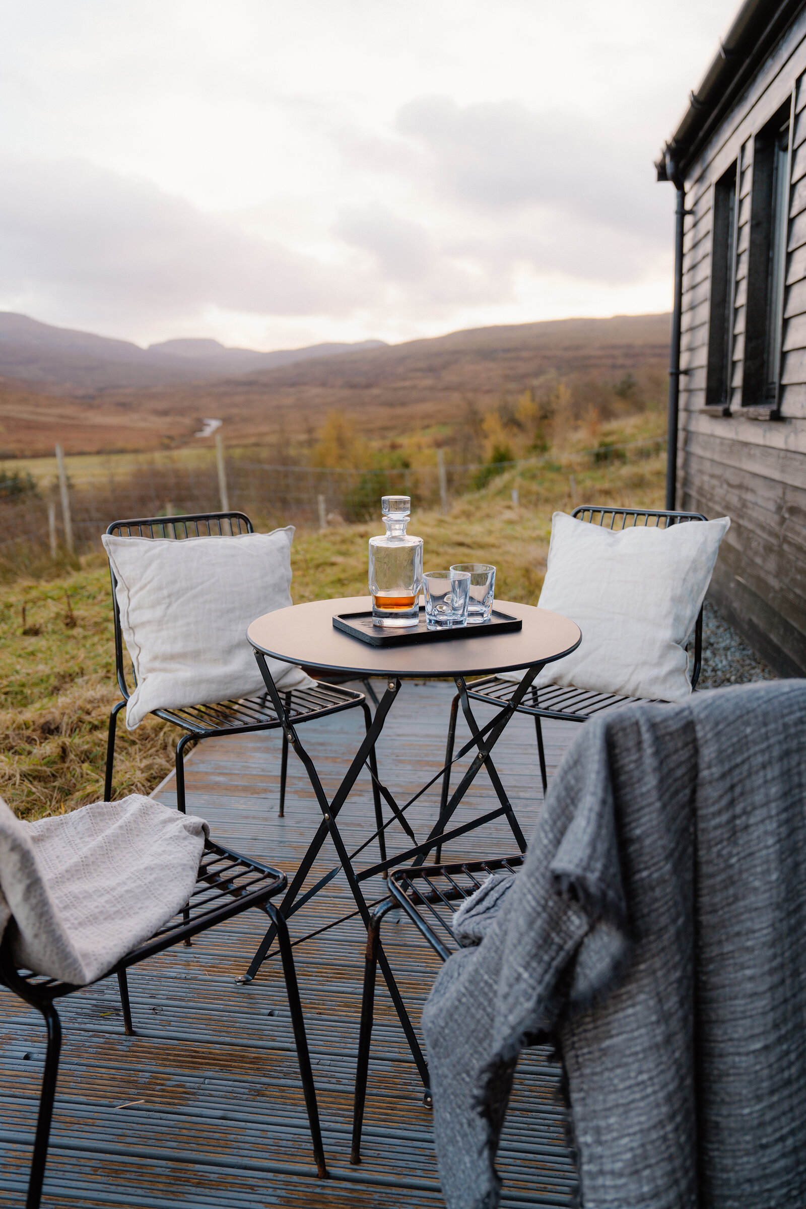 A whisky decanter and glasses sit on a patio table overlooking moorland hills.
