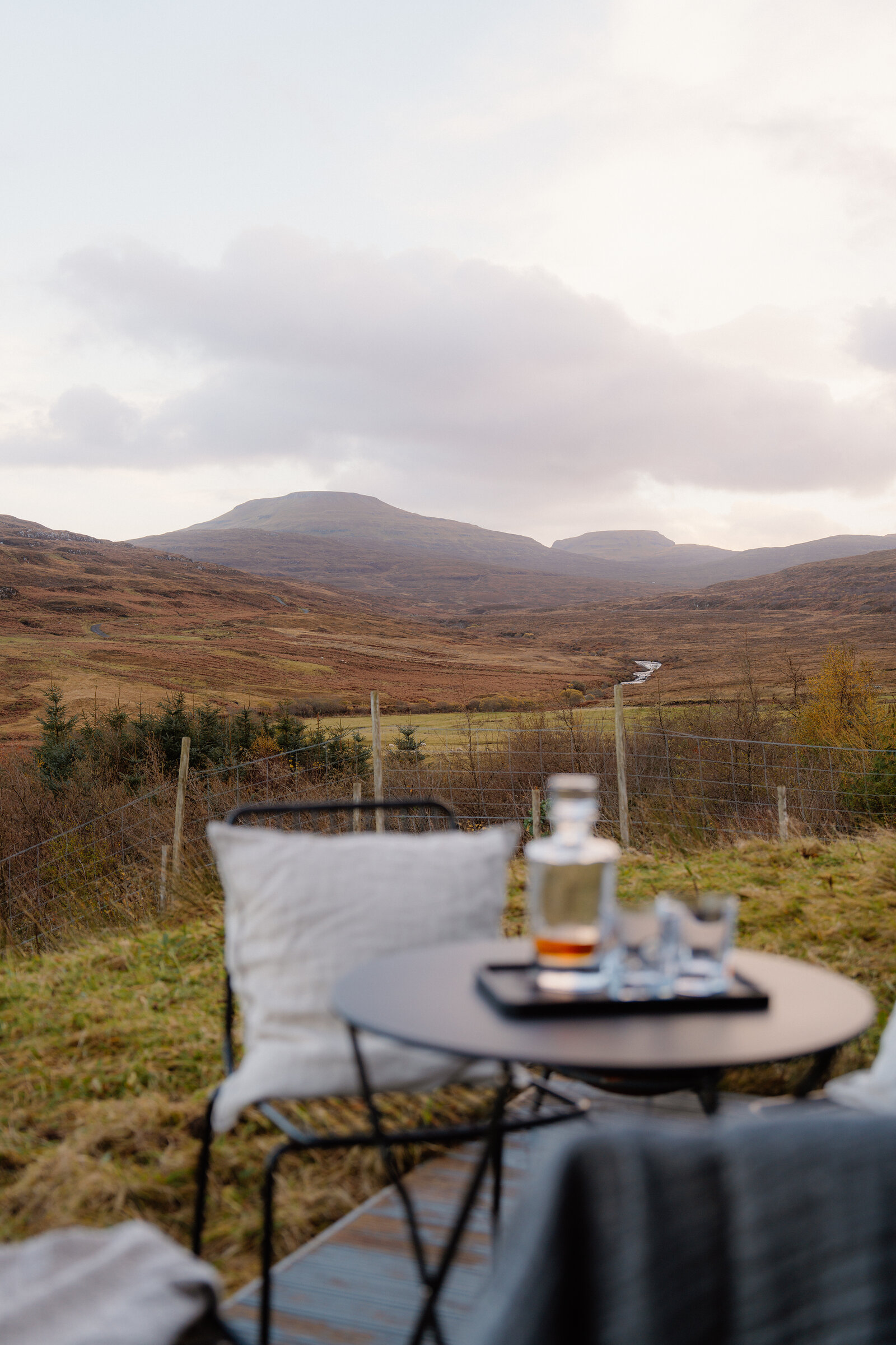 A whisky decanter and glasses sit on a patio table before distant hills.