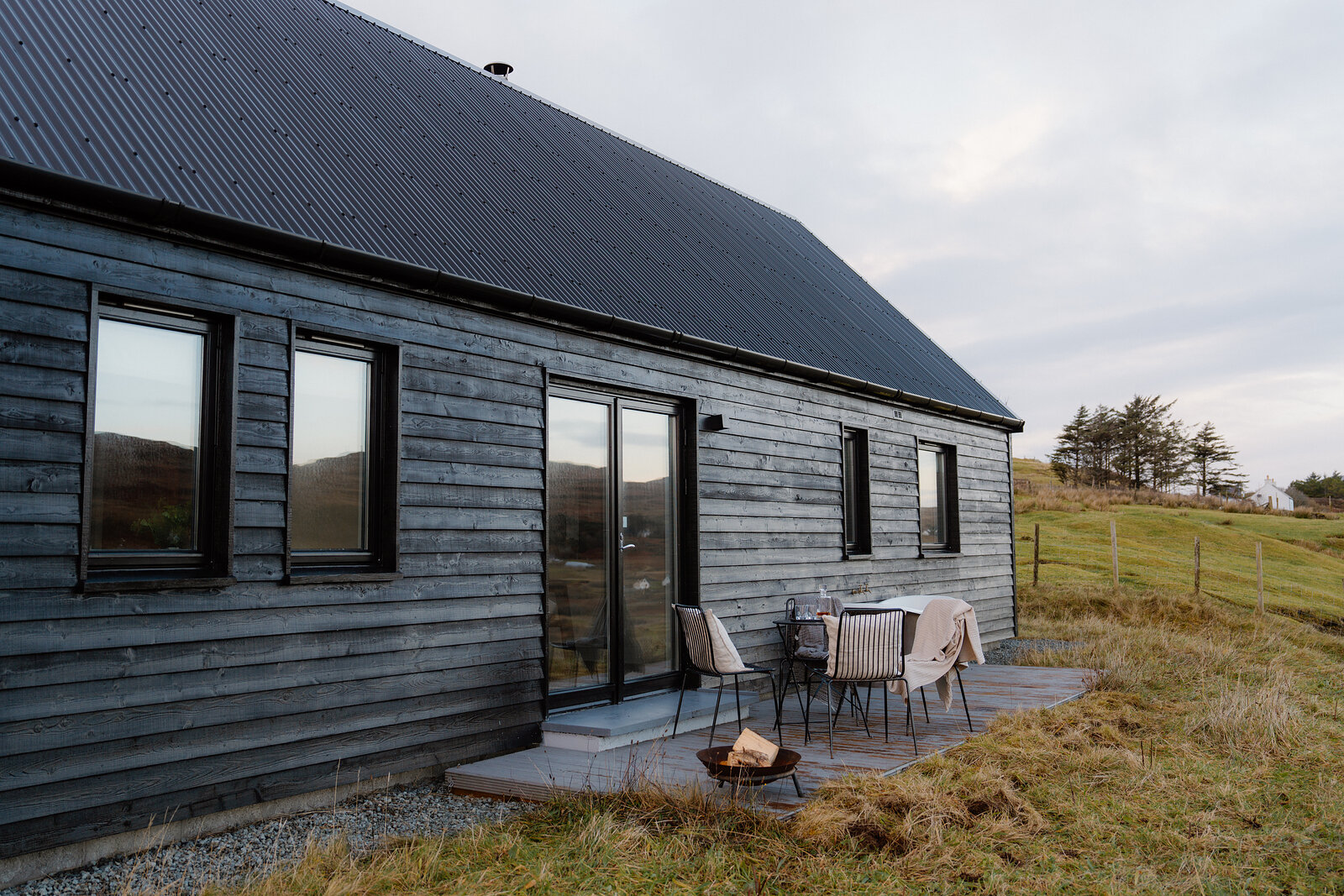 Outdoor chairs and a fire pit sit on a patio beside a black timber-clad house.