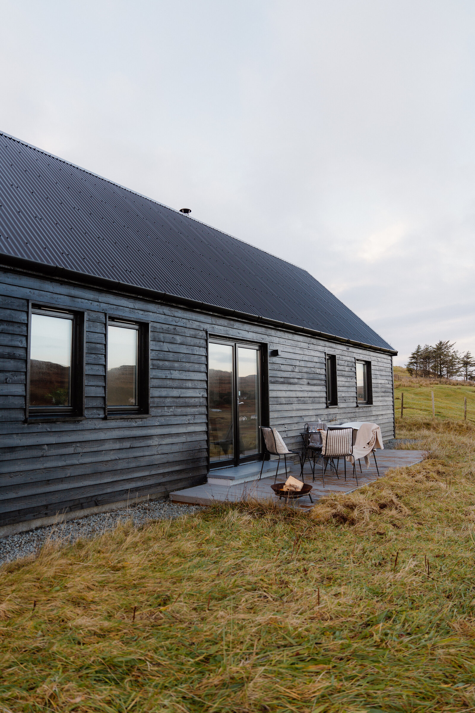 Chairs and a fire pit sit on a patio beside a black timber-clad house at dusk.