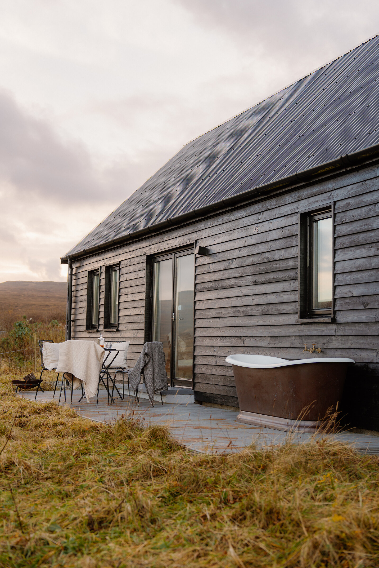 A freestanding copper bathtub sits on a deck beside a black timber-clad house.