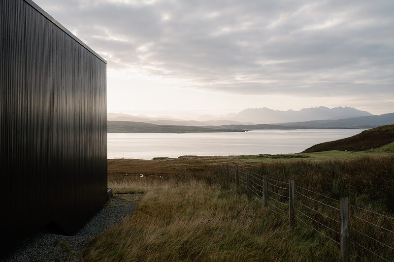 A black timber house stands before a loch with distant jagged mountains.