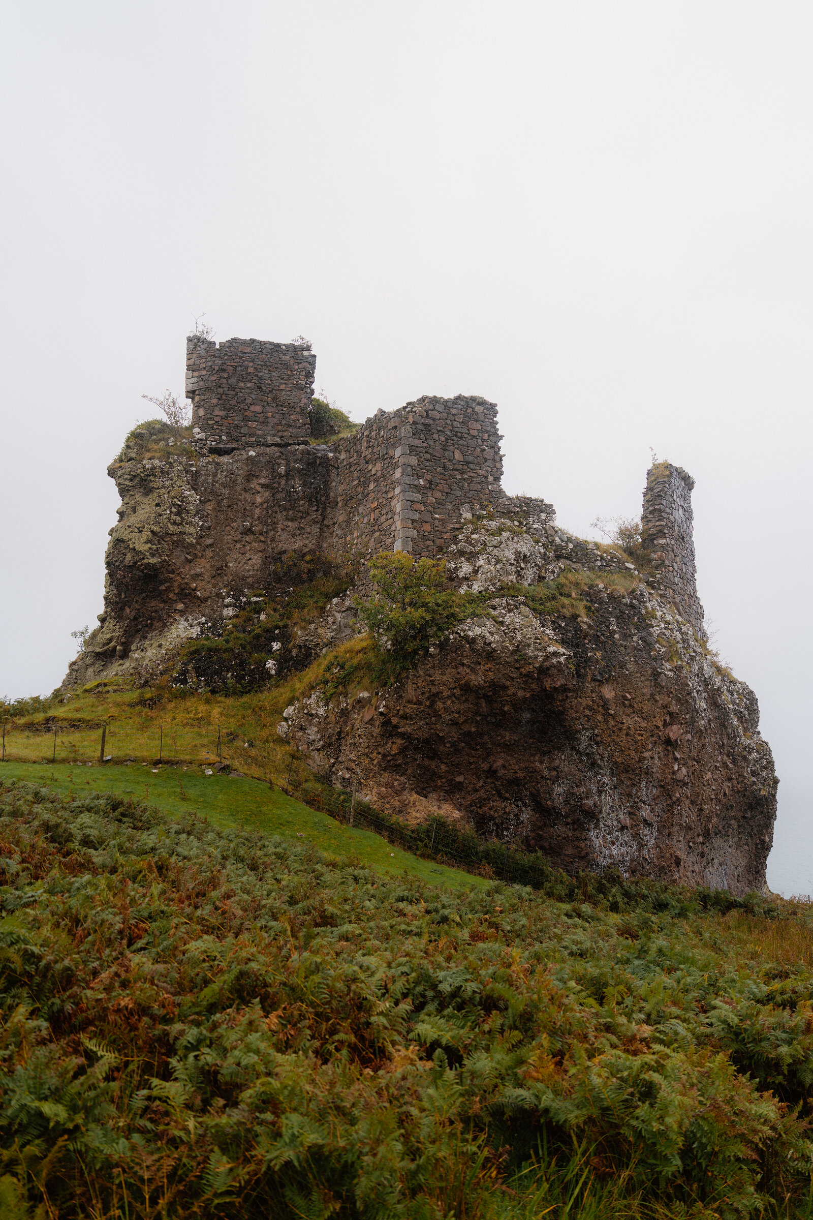 A ruined stone castle sits on a rocky crag above green and rust-coloured hillside.