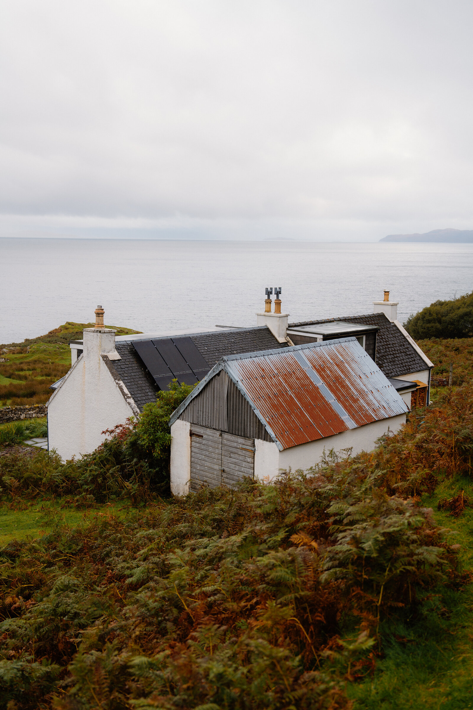 A white cottage with solar panels stands beside a rusted corrugated outbuilding on a coastal hillside.