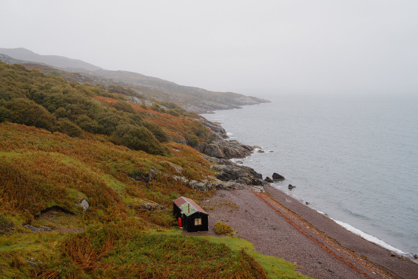 A small hut sits alone on a shingle beach below a misty, heather-covered coastal hillside.