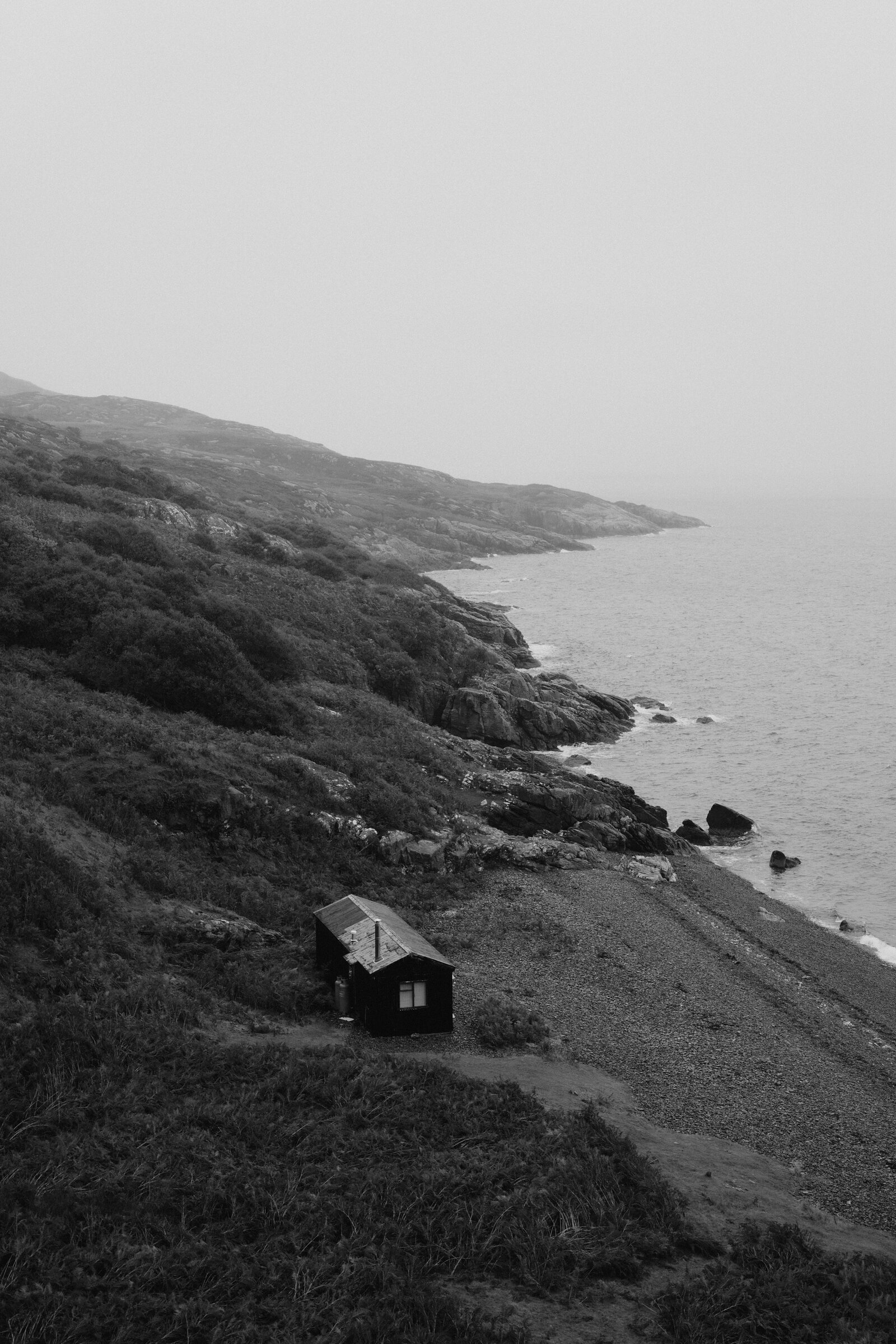 A black-and-white view shows a small hut on a shingle beach below a misty coastal hillside.