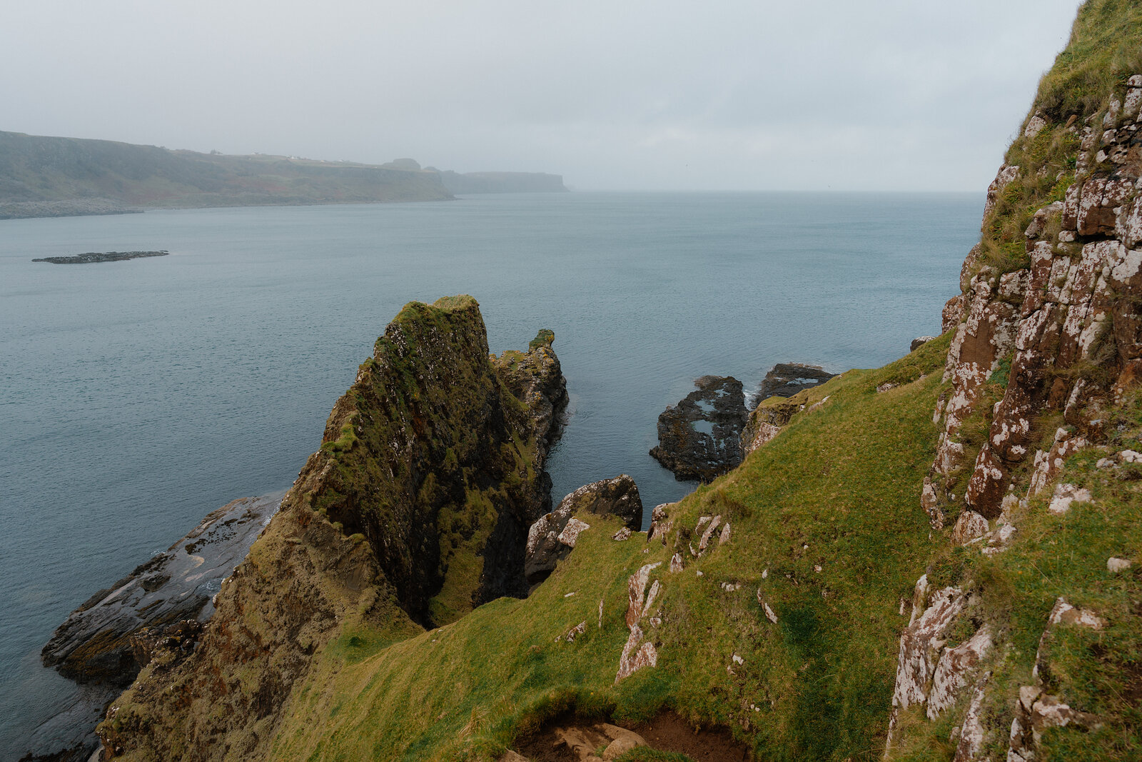A jagged rock stack divides calm water beside a misty coastline.