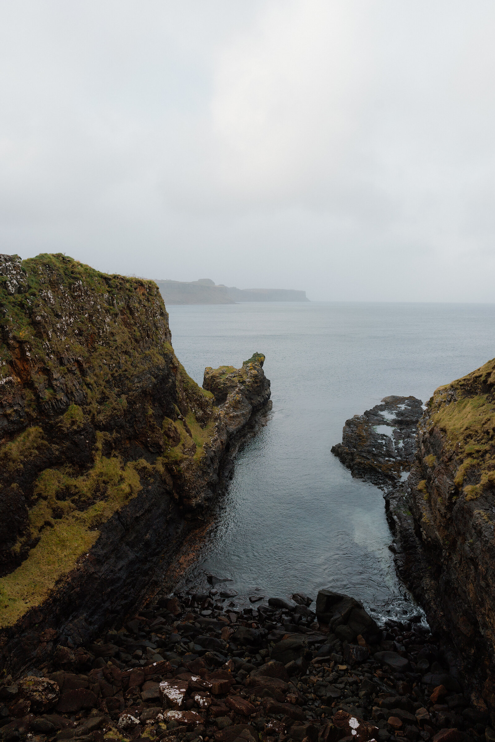 A narrow sea inlet cuts between mossy rock walls beneath a misty headland.