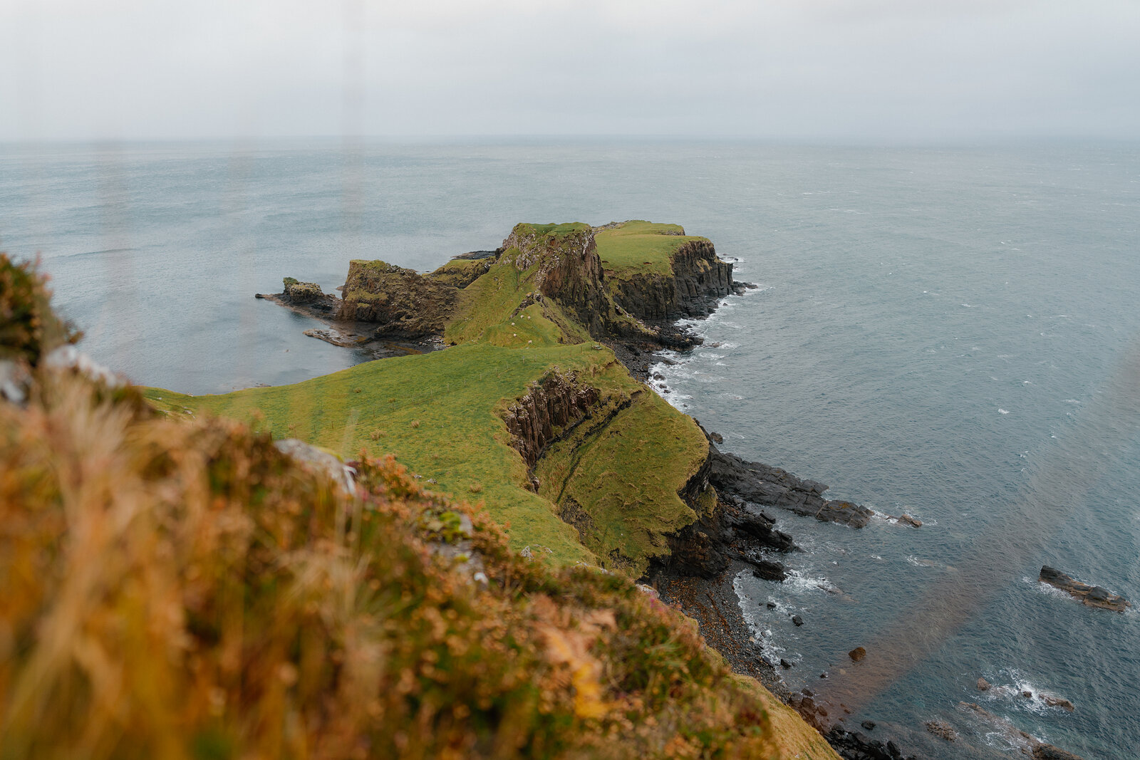 A grassy headland with a saddle-shaped ridge extends into the sea beneath overcast skies.