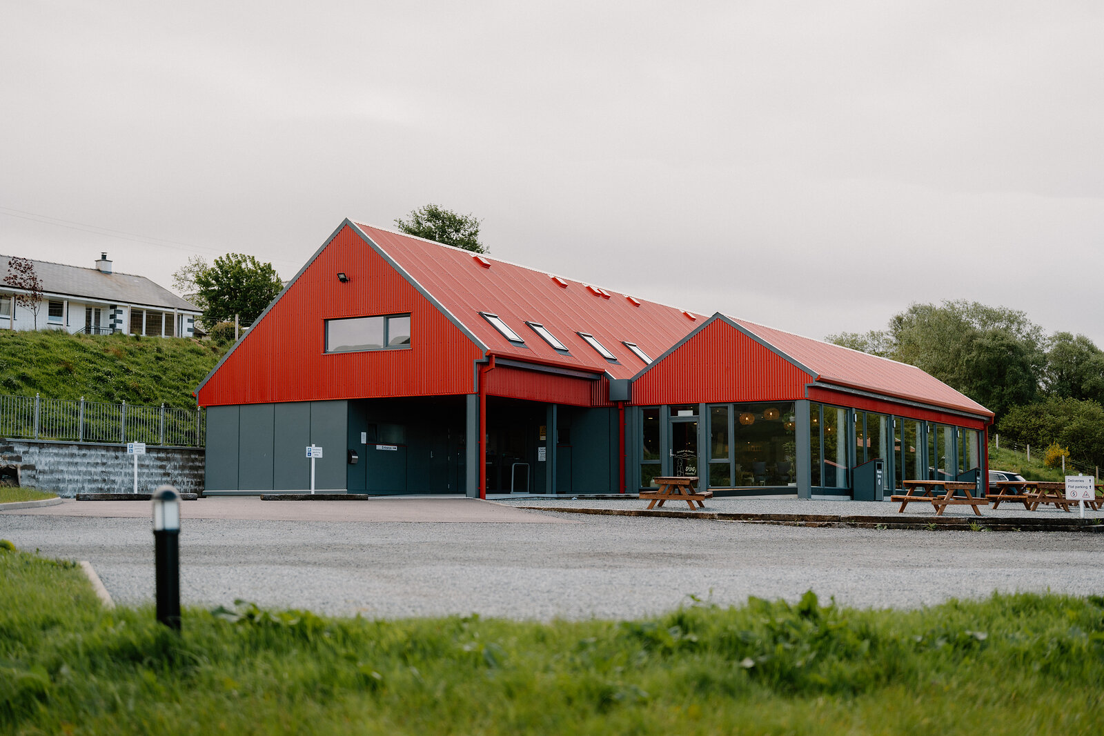 A red-clad gabled building with grey trim stands beside a car park and picnic tables.