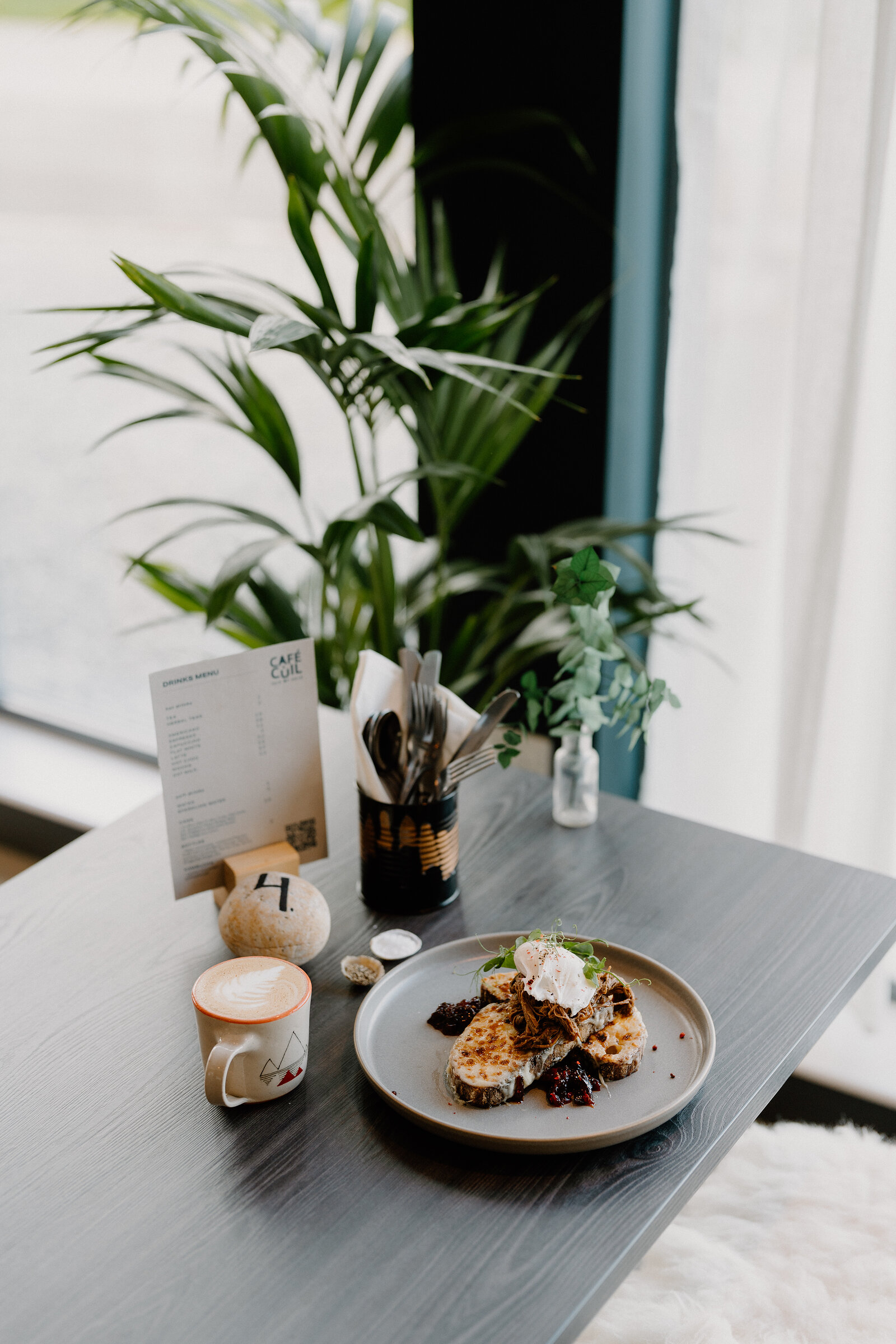 A Café Cùil menu card stands beside a latte and a plated dish of toast, pulled meat and poached egg.