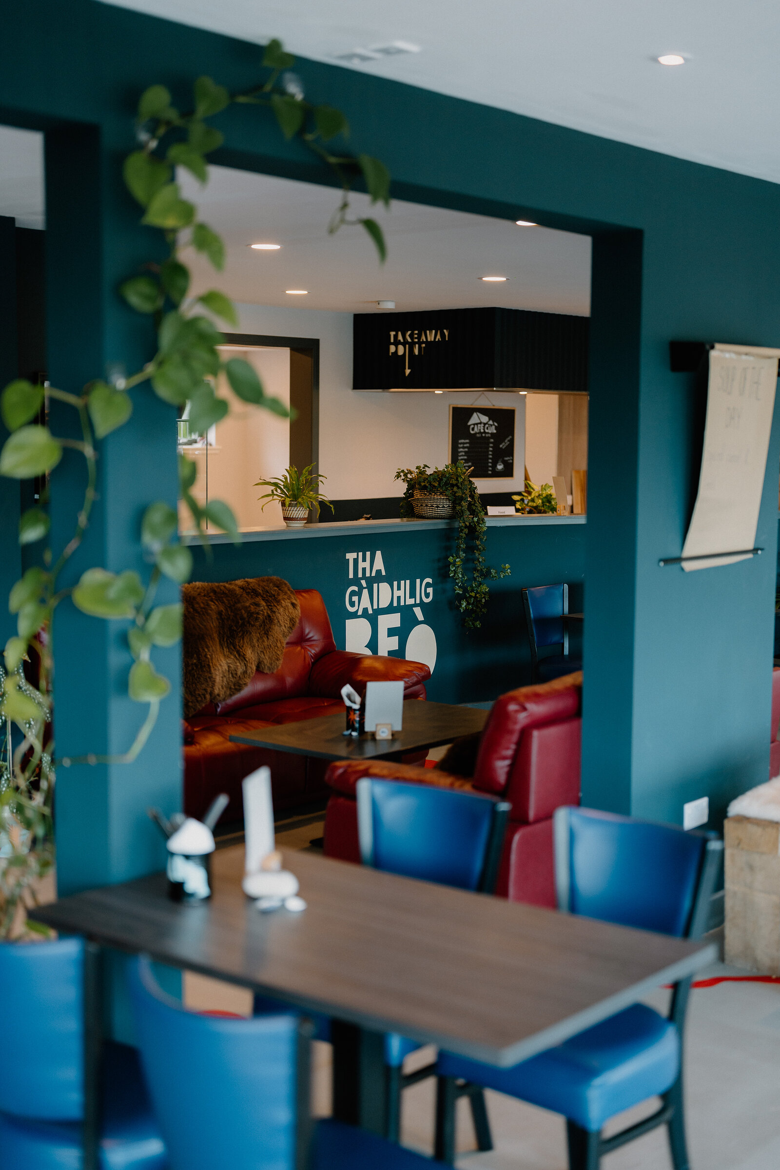 A wall reads 'Tha Gàidhlig Beò' above a red leather sofa in a teal-walled cafe lounge.