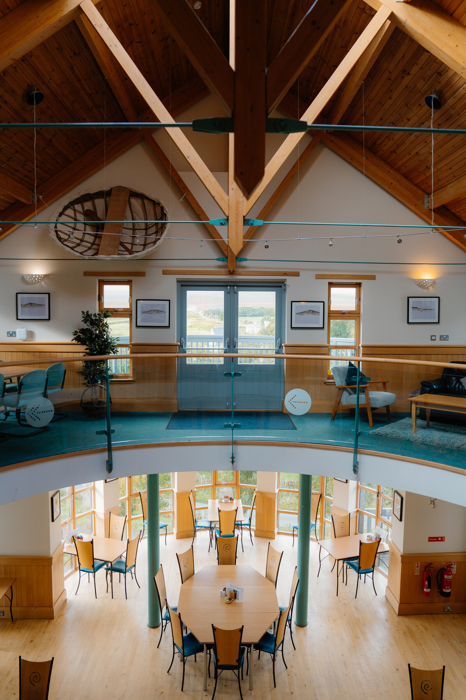 Wooden tables and chairs fill a circular dining hall beneath a timber-raftered upper gallery.