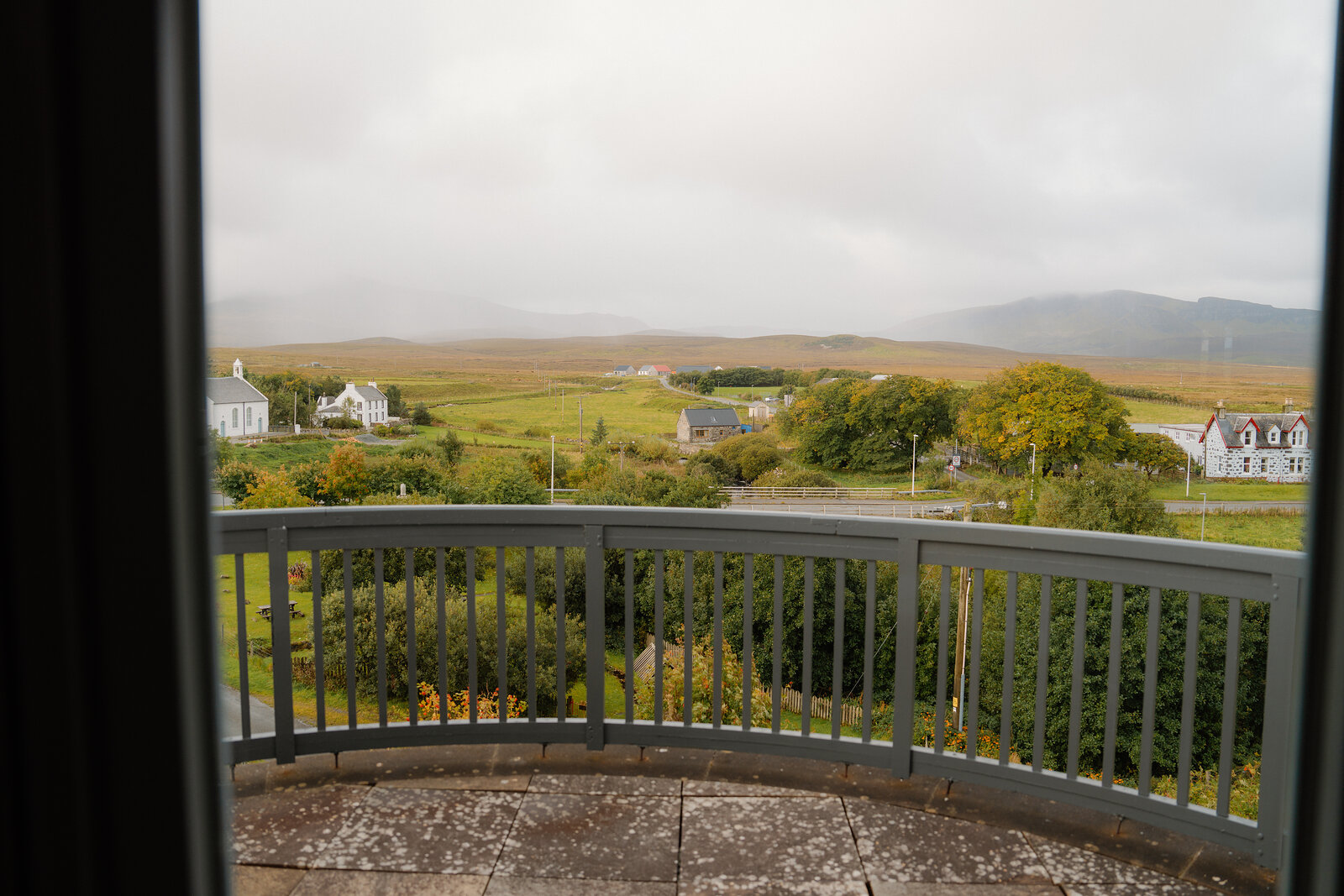 A curved balcony overlooks a misty village and moorland hills.