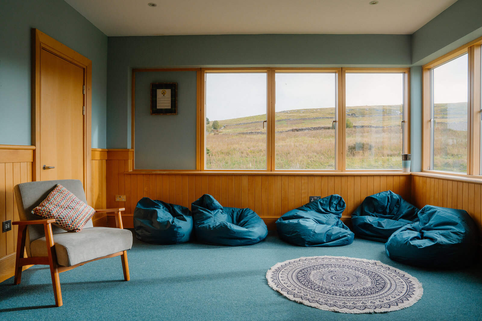 Blue beanbags line a carpeted room with a round rug, overlooking moorland through large windows.