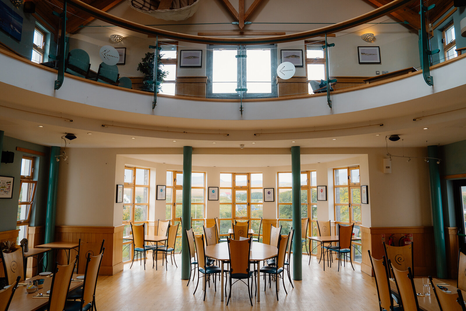 Round tables and chairs are arranged beneath a mezzanine gallery in a sunlit circular hall.