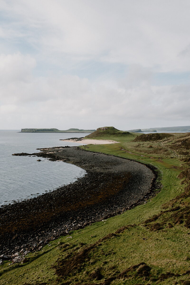 A sandy tombolo curves beneath a grassy headland with a small island visible offshore.