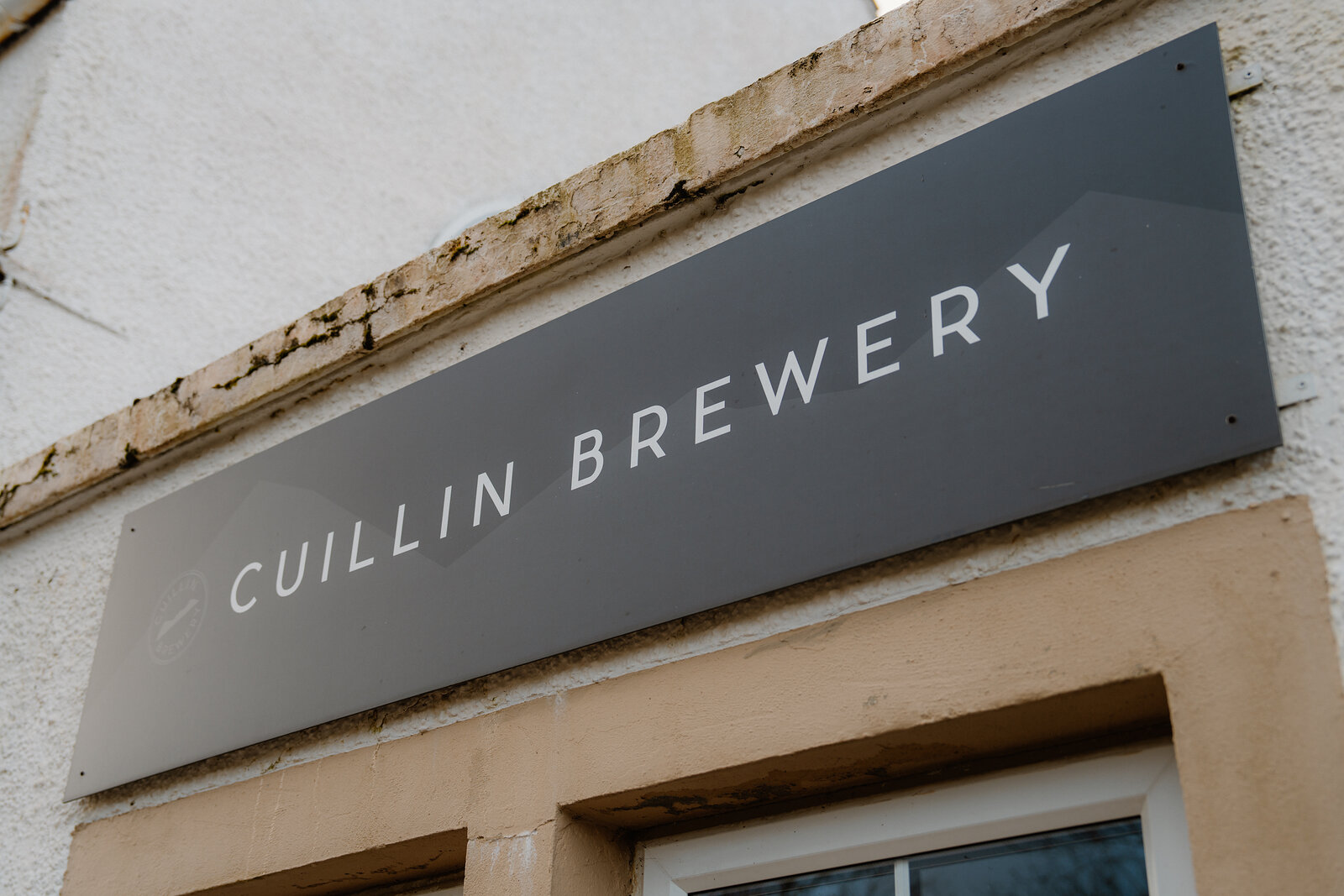A grey sign reading 'Cuillin Brewery' is mounted above a doorway on a whitewashed building on Skye.