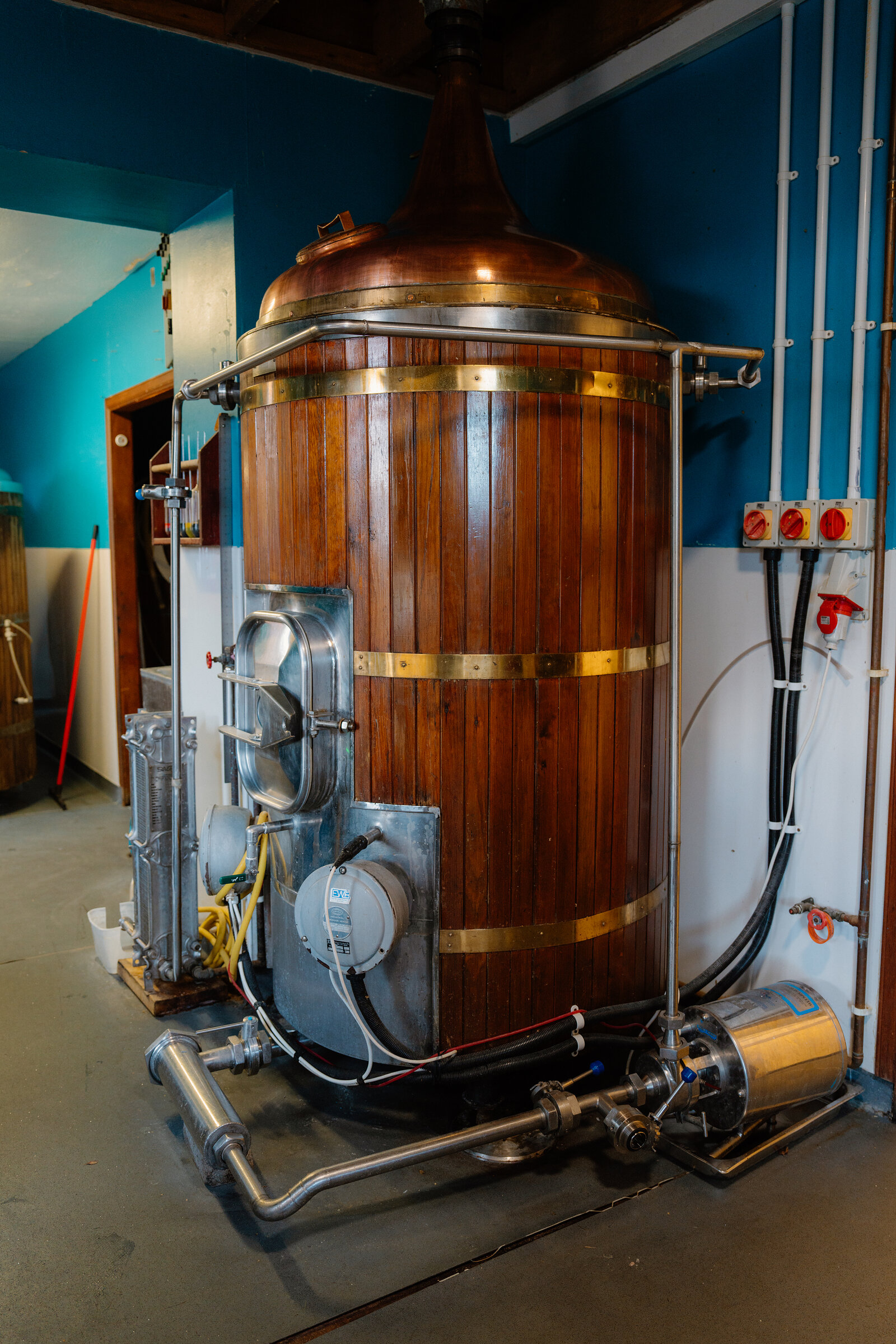 A copper-topped, wood-clad brewing kettle stands fitted with gauges and pipework in a blue-walled brewery.