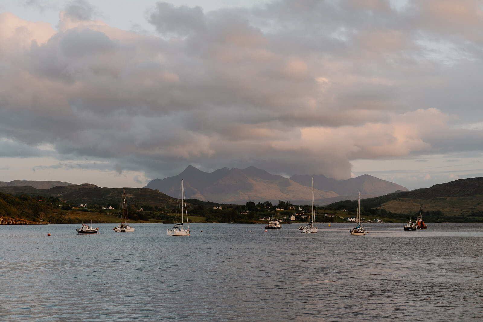 Sailboats are moored in a bay beneath the jagged silhouette of the Cuillin mountains at dusk.