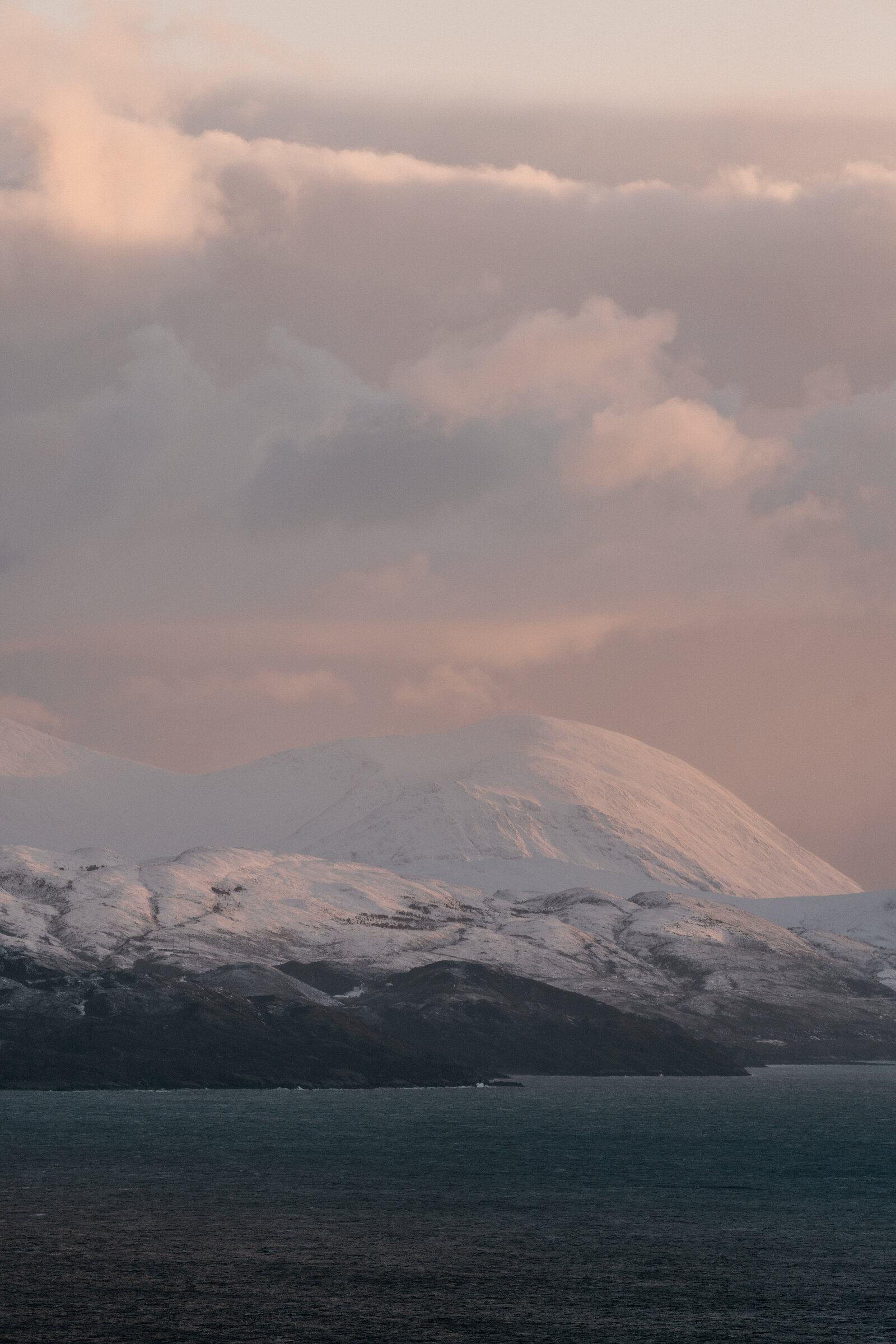 A snow-capped mountain rises above a dark coastline beneath a pink evening sky.