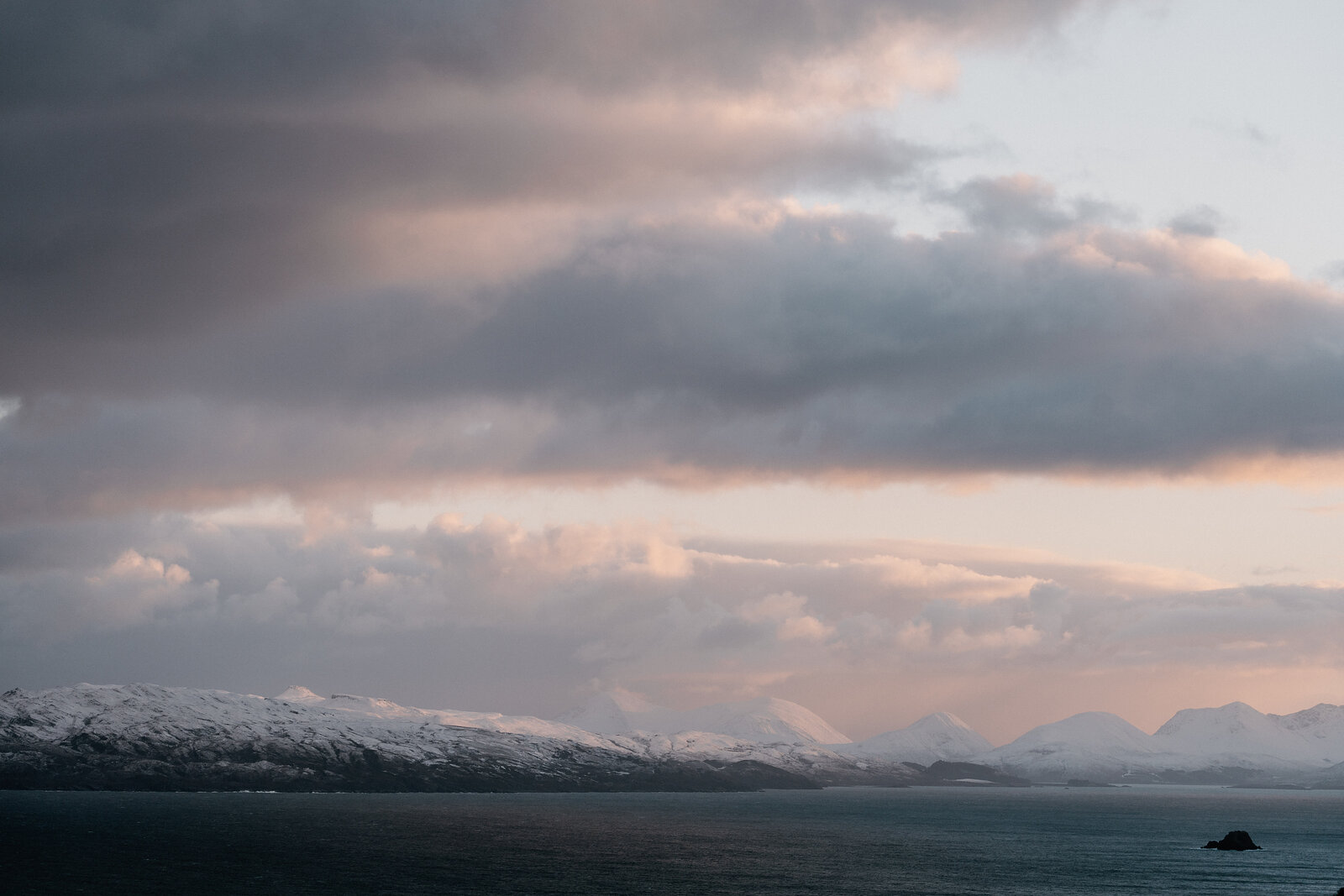 Pink-tinged clouds stretch above snow-capped mountains along a dark shoreline.