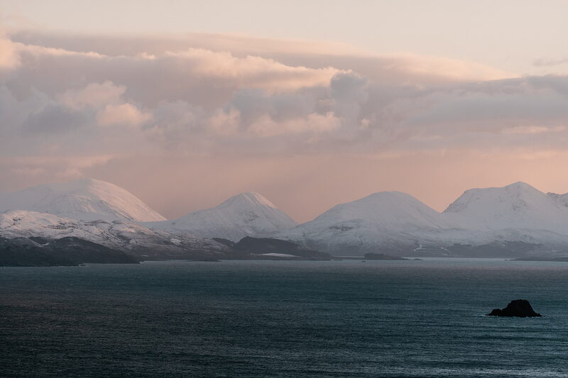 Snow-capped peaks rise along a coastline beneath pink evening clouds, a small rocky islet in the foreground sea.