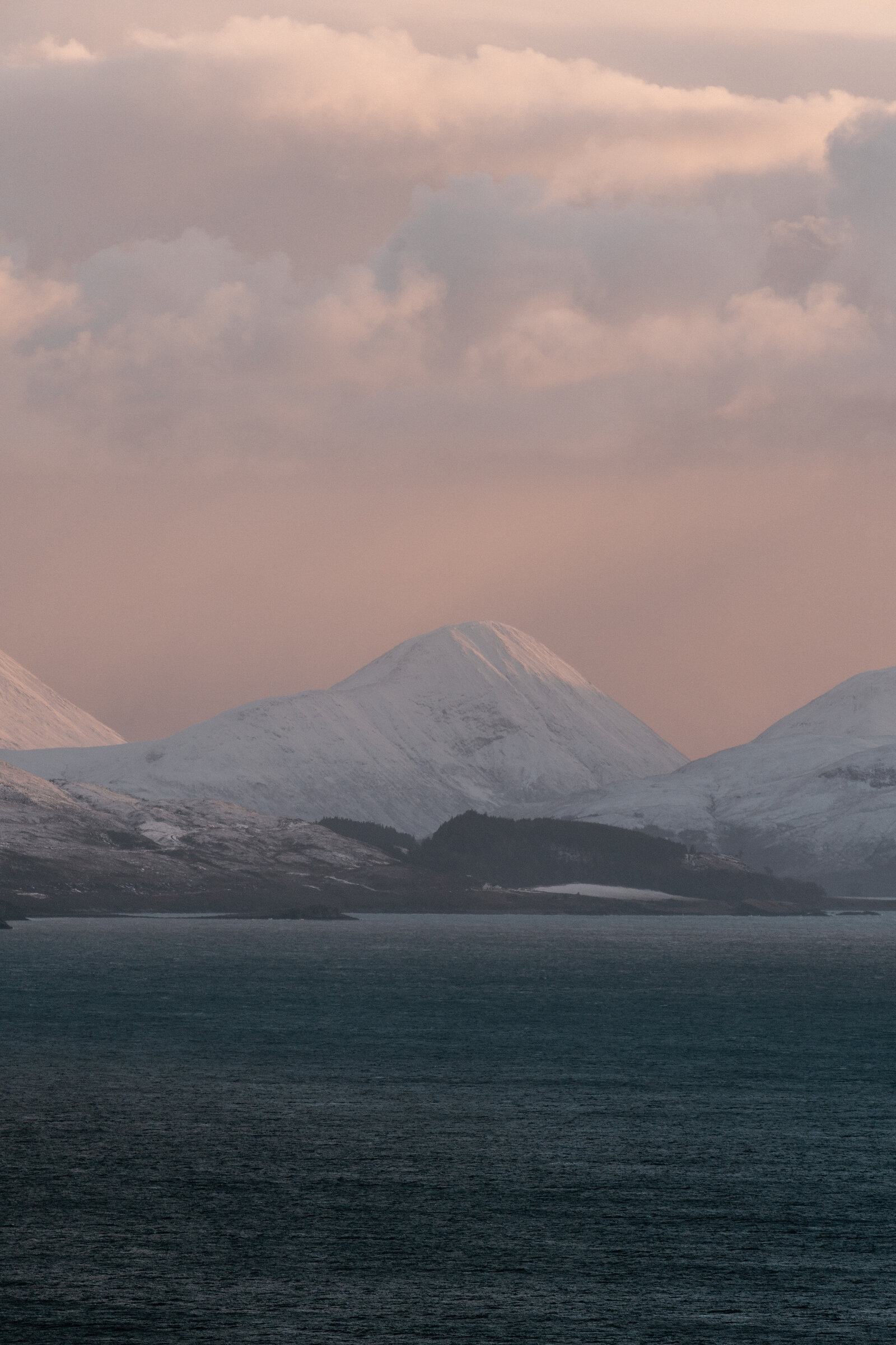 A snow-capped peak rises above a wooded shoreline beneath a hazy pink sky.